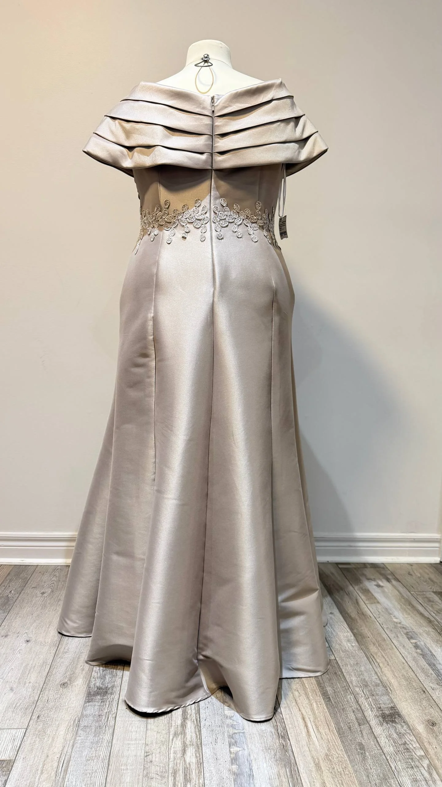 A wedding dress displayed on a mannequin with off-the-shoulder layered ruffle details, embroidered floral appliqué around the waist, and a satin finish in a light champagne color.