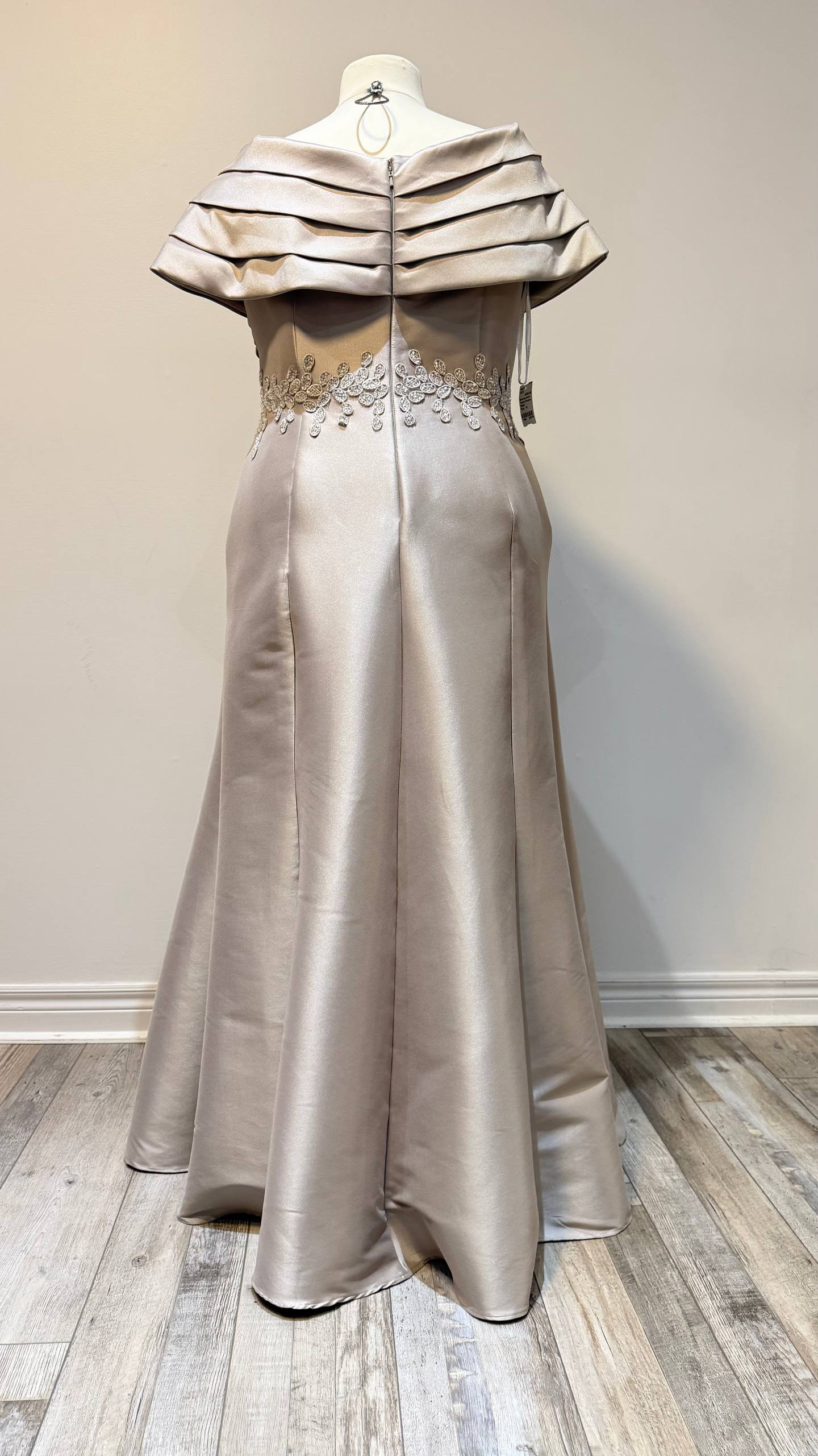 A strapless satin wedding dress on a mannequin. The dress features intricate embroidery at the waist and a tiered ruffled off-shoulder overlay on the top. The background is a plain beige wall, and the floor is wooden.