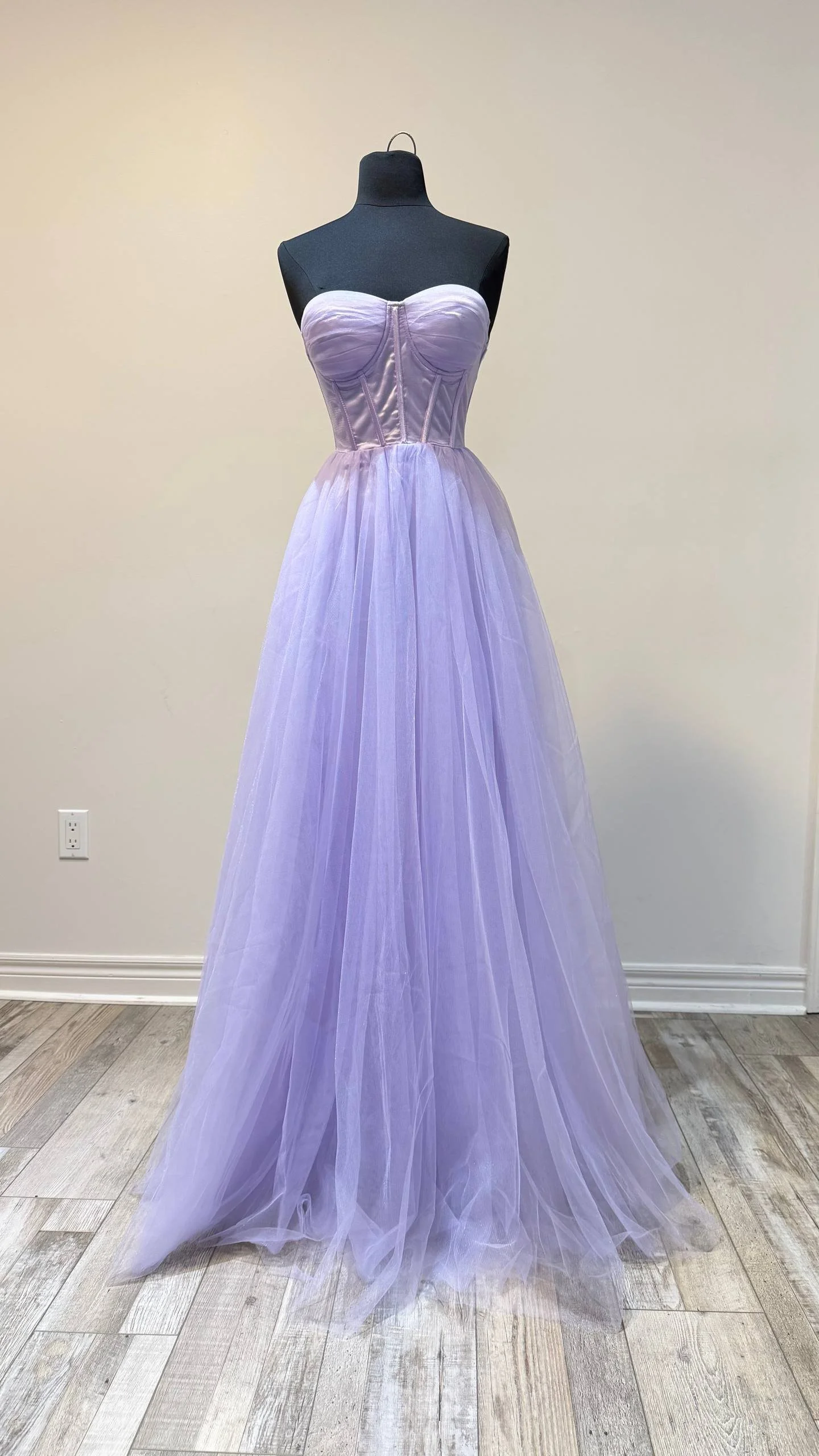 A lavender strapless gown with a fitted bodice and full tulle skirt displayed on a black mannequin against a plain beige wall.