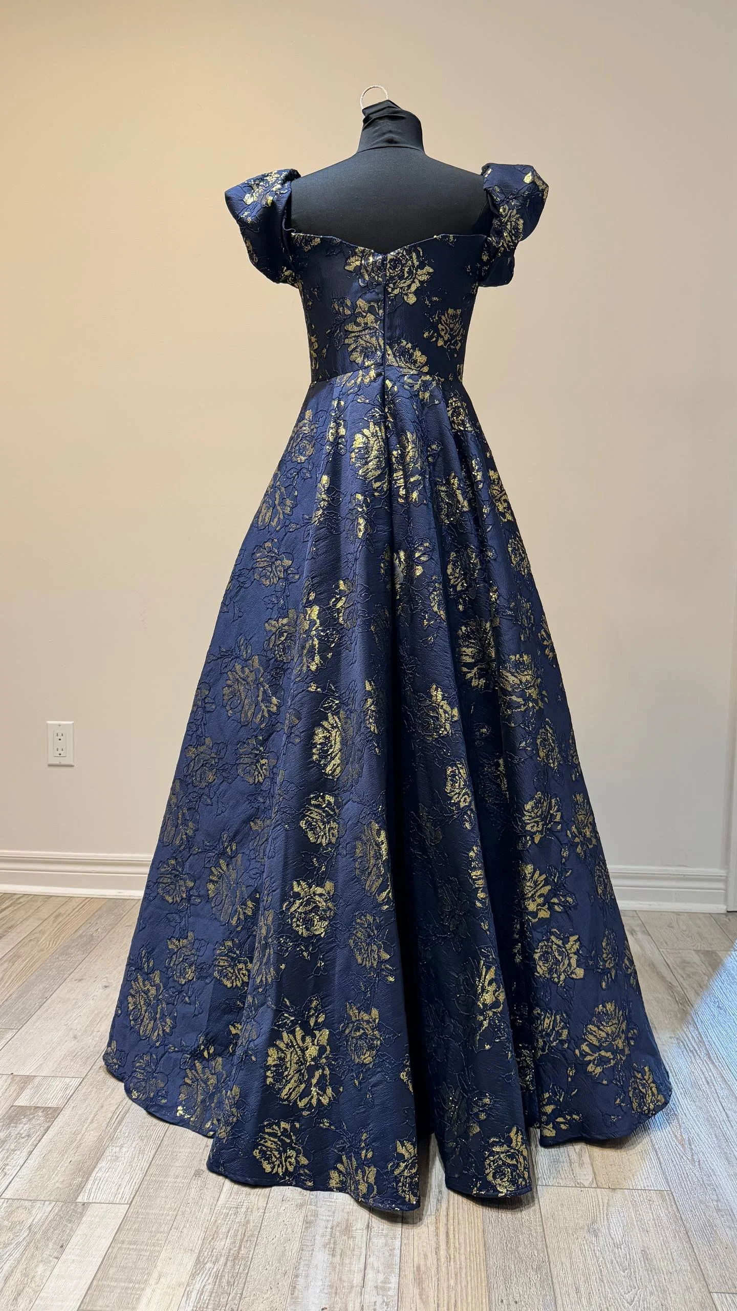 A navy blue evening gown with gold floral embroidery displayed on a mannequin. The dress has a fitted bodice, puffed short sleeves, and a long, flowing skirt.