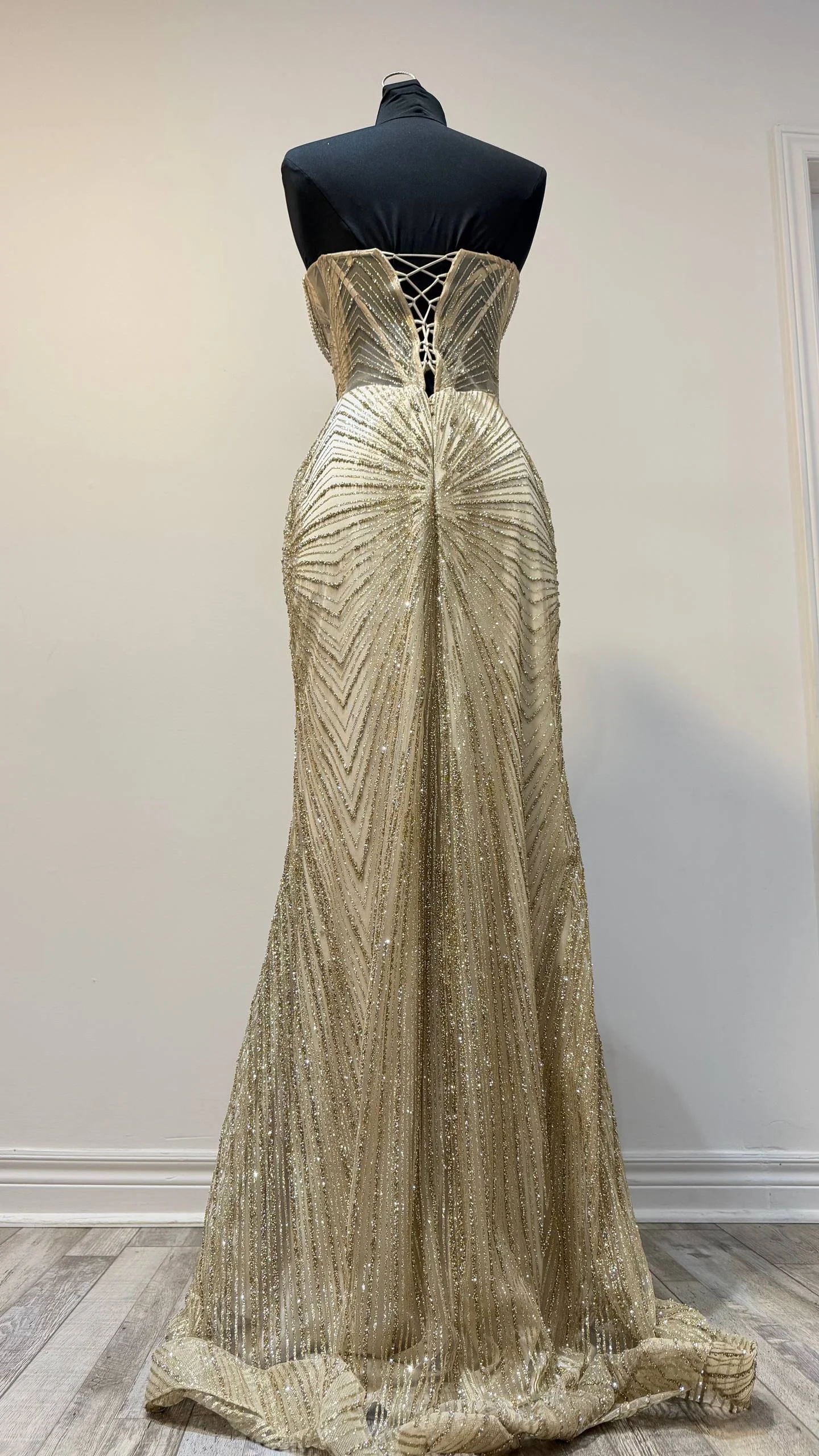 A gold, sequined evening gown displayed on a black mannequin against a white wall.