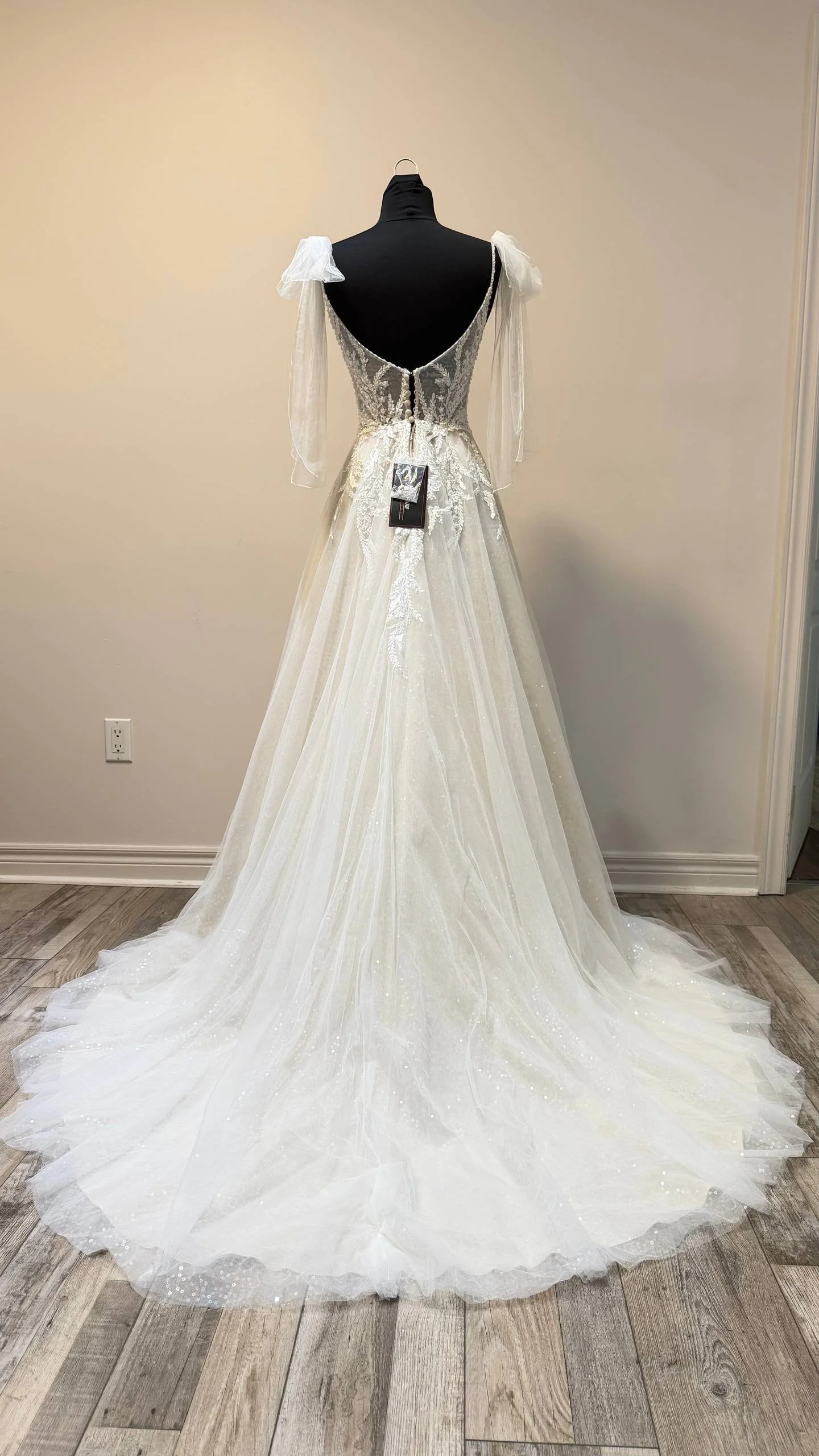 A wedding dress displayed on a mannequin, featuring a fitted bodice with lace details, sheer long sleeves, and a flowing train with a soft shimmer, placed against a plain beige wall.