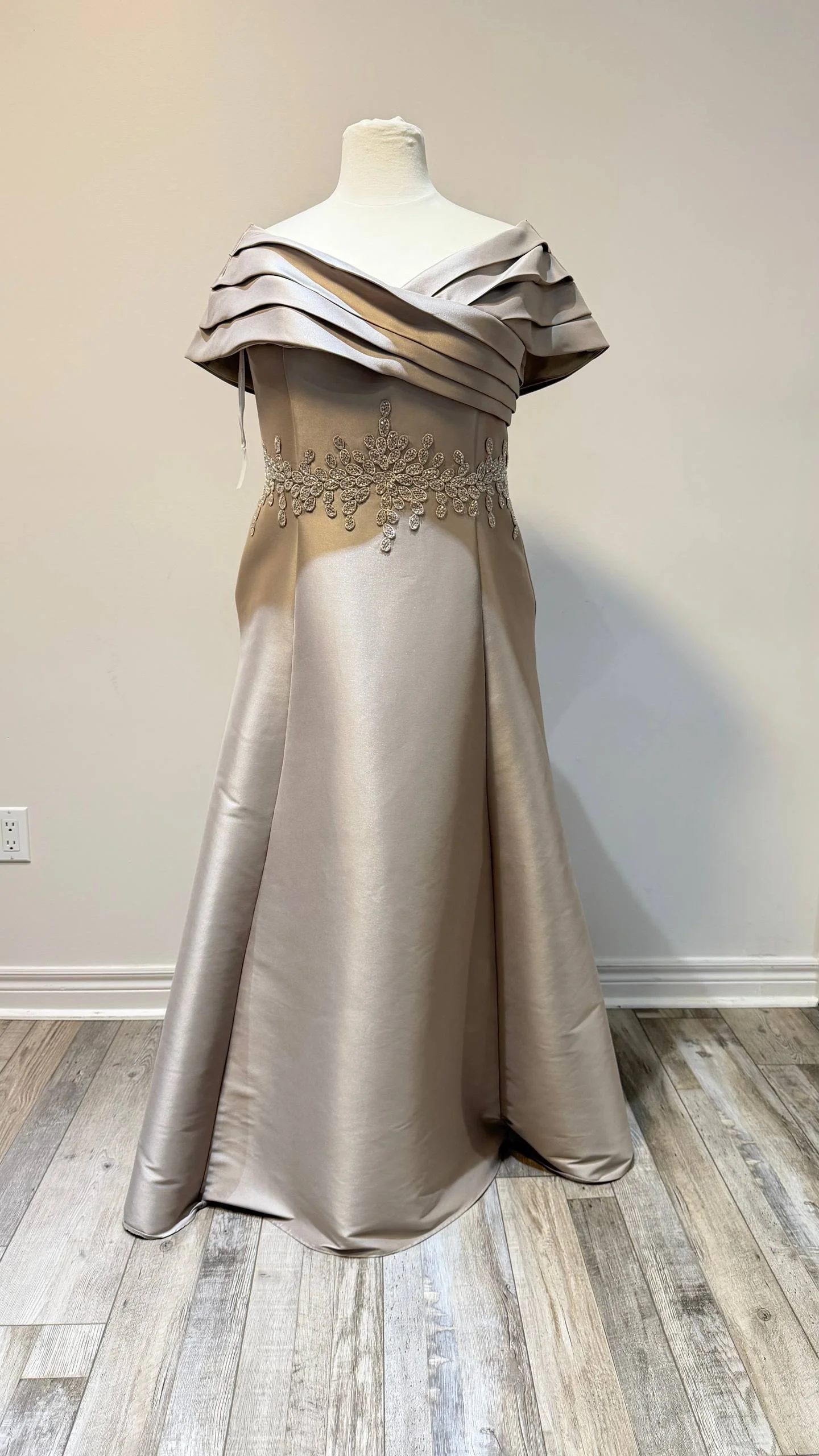 A formal evening gown on a mannequin with a satin finish, featuring an off-shoulder ruffled top and intricate beaded floral embroidery at the waist.