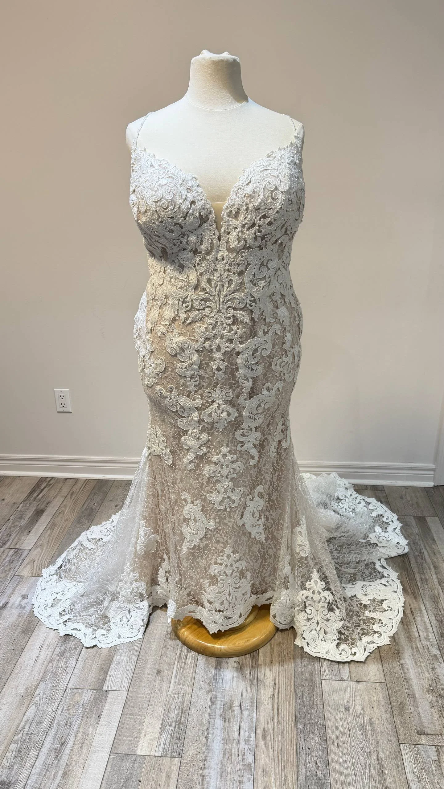 A white lace wedding dress displayed on a mannequin with a clear neckline and intricate floral lace patterns, fitted silhouette, and train on a wooden floor.