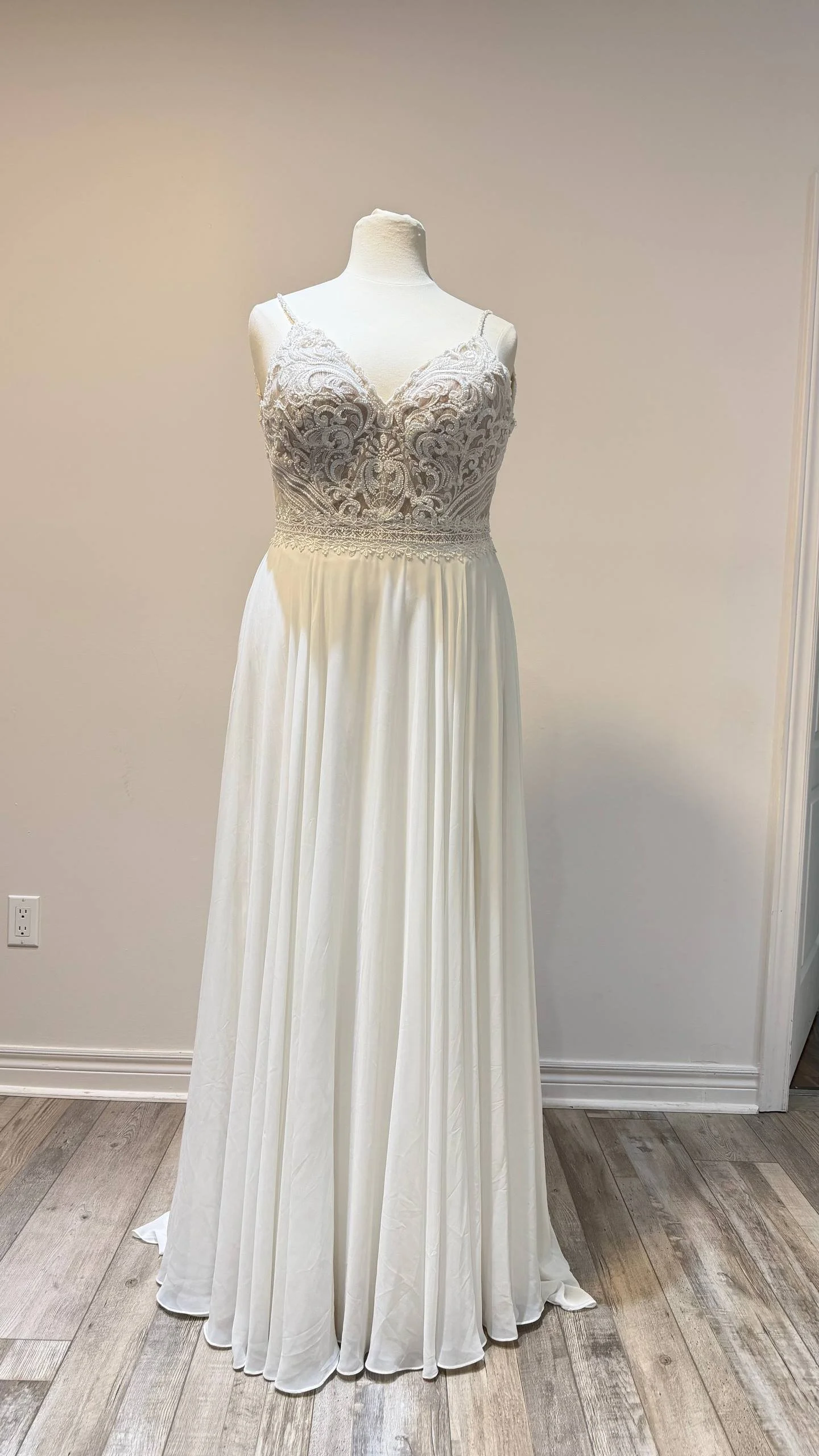 White wedding dress displayed on a mannequin with intricate lace bodice and flowing skirt, set against a plain wall.