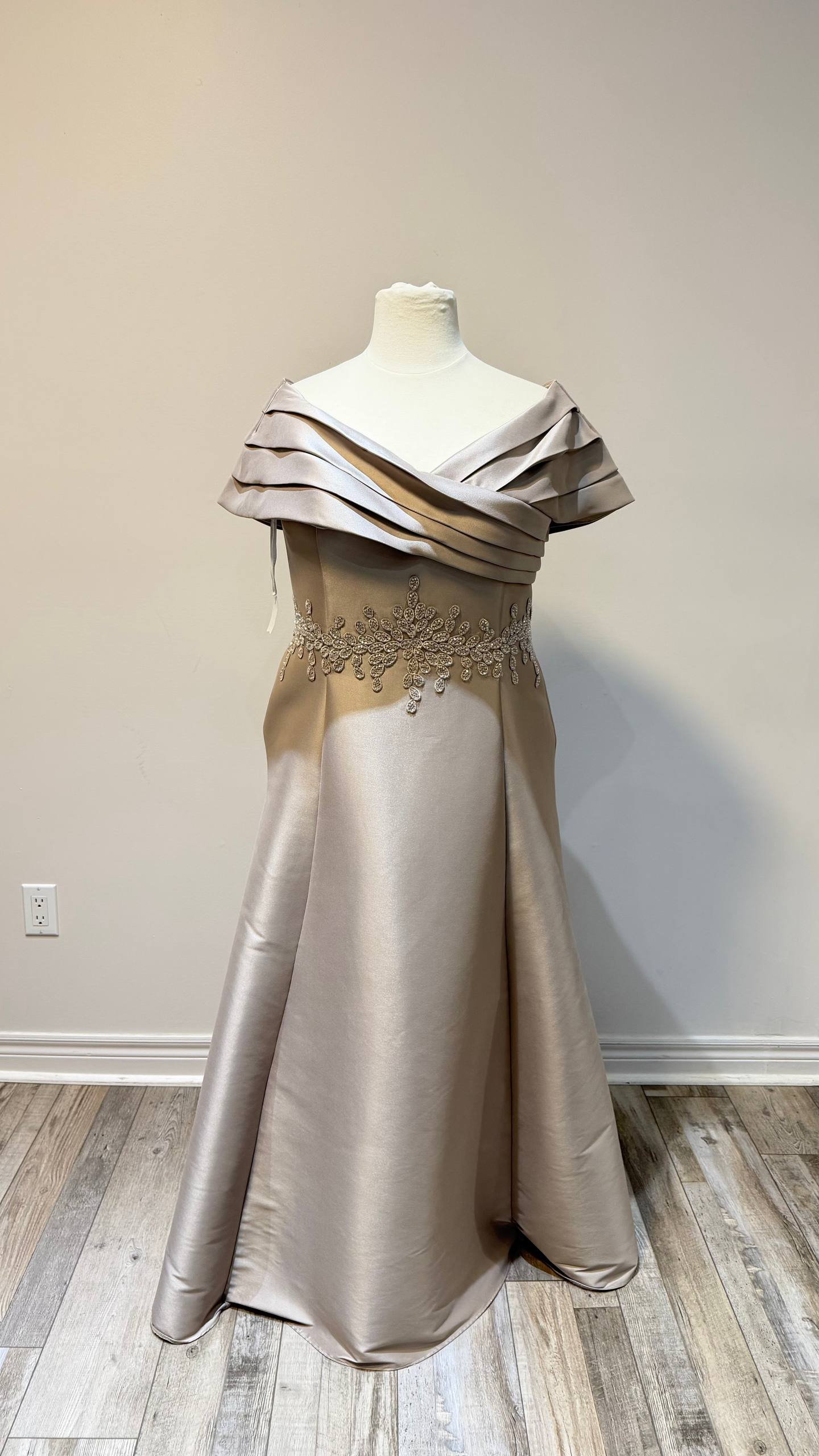 A beige satin gown featuring off-the-shoulder layered ruffle sleeves and intricate floral embellishments around the waist, displayed on a mannequin against a plain wall.