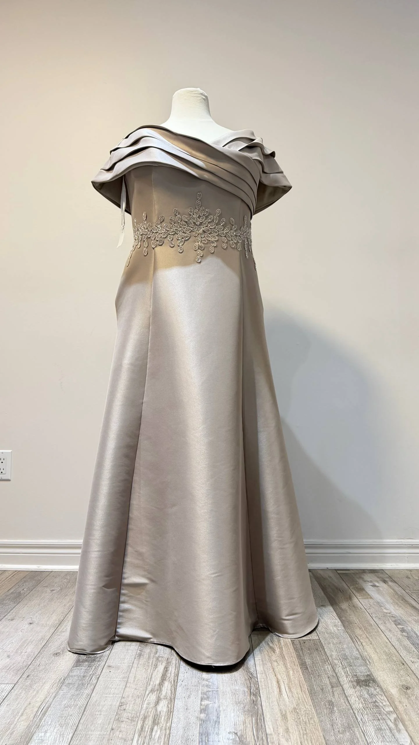 A formal gown on a dress form featuring a satin fabric with a fitted bodice adorned with intricate beadwork and a wide, ruffled off-shoulder style neckline.