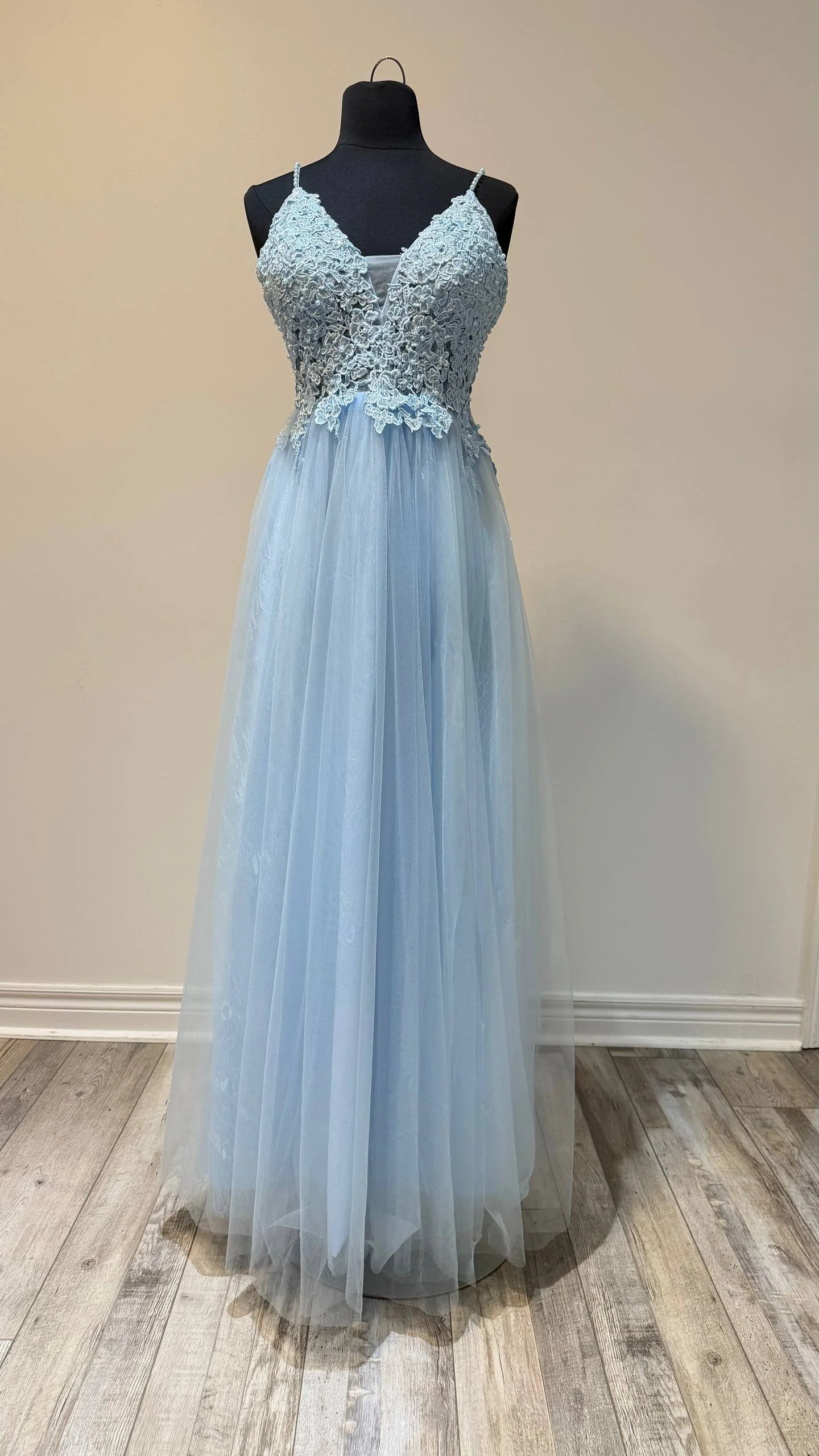 A light blue, sleeveless evening gown with lace bodice and flowy tulle skirt displayed on a black dress form against a beige wall.