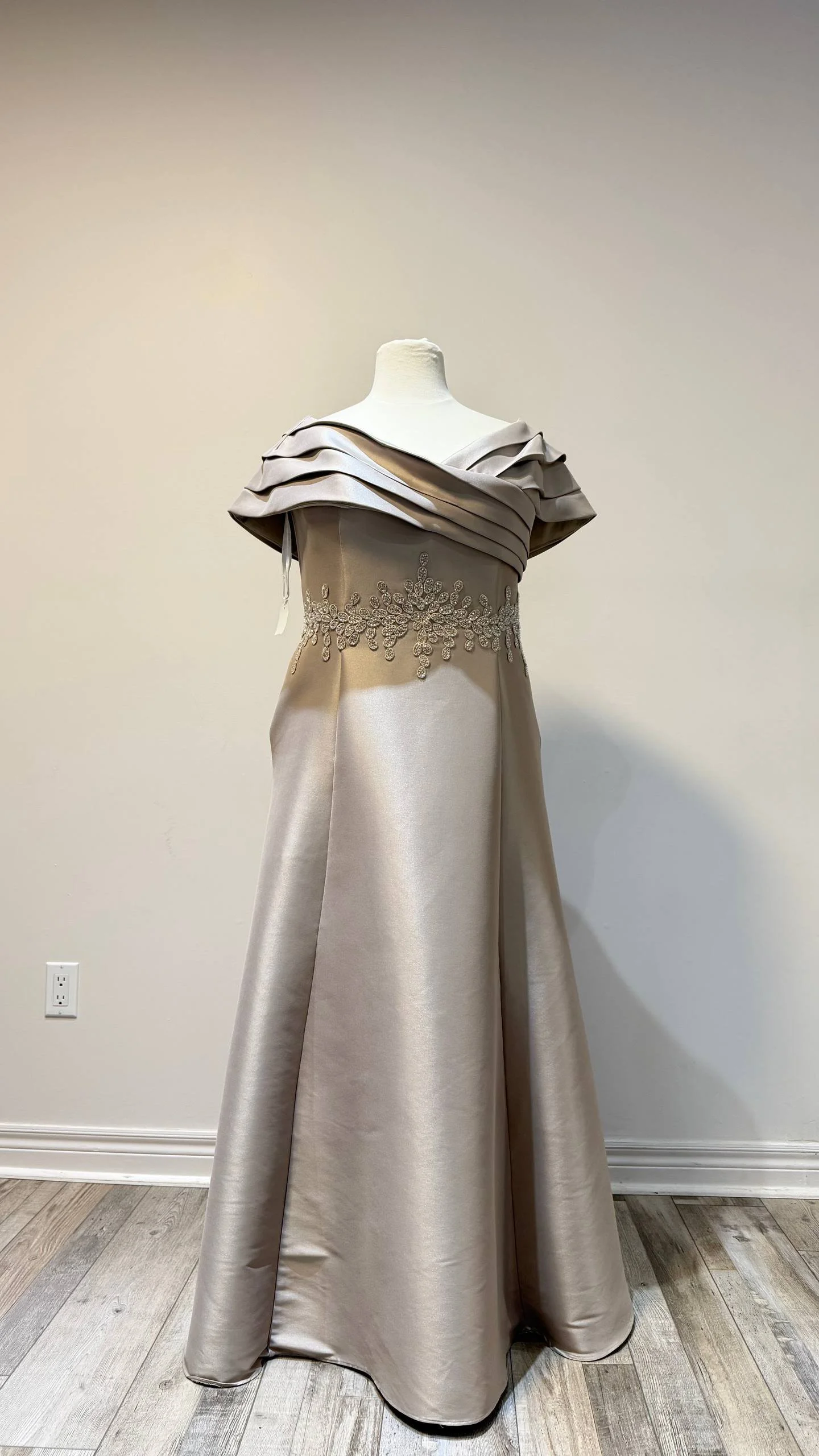 A beige satin evening gown with an off-the-shoulder design and draped fabric at the bodice, decorated with intricate beadwork and embellishments.