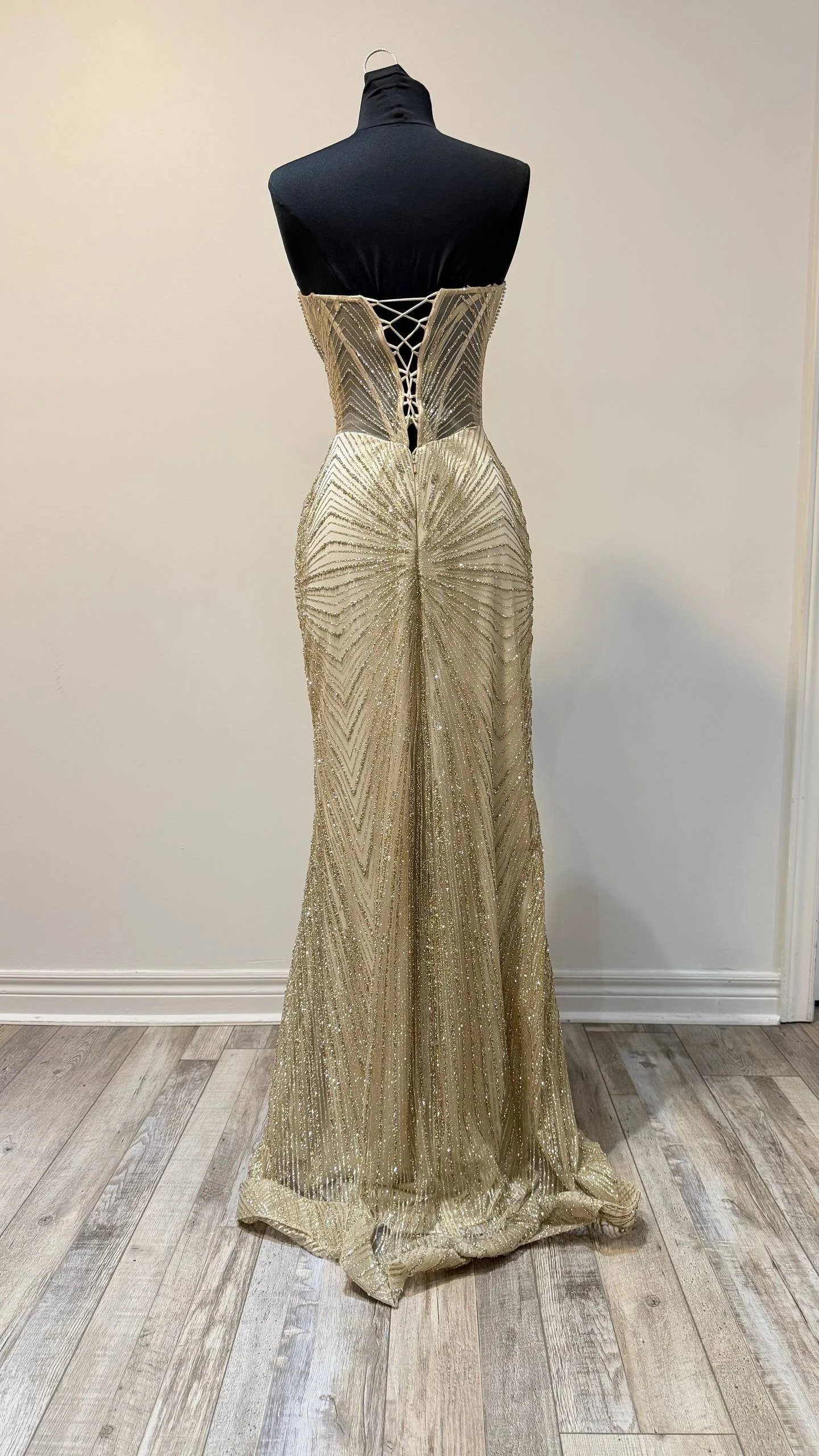A gold, glittery evening gown on a mannequin with a black top and high neck, displayed against a plain wall and wooden floor.
