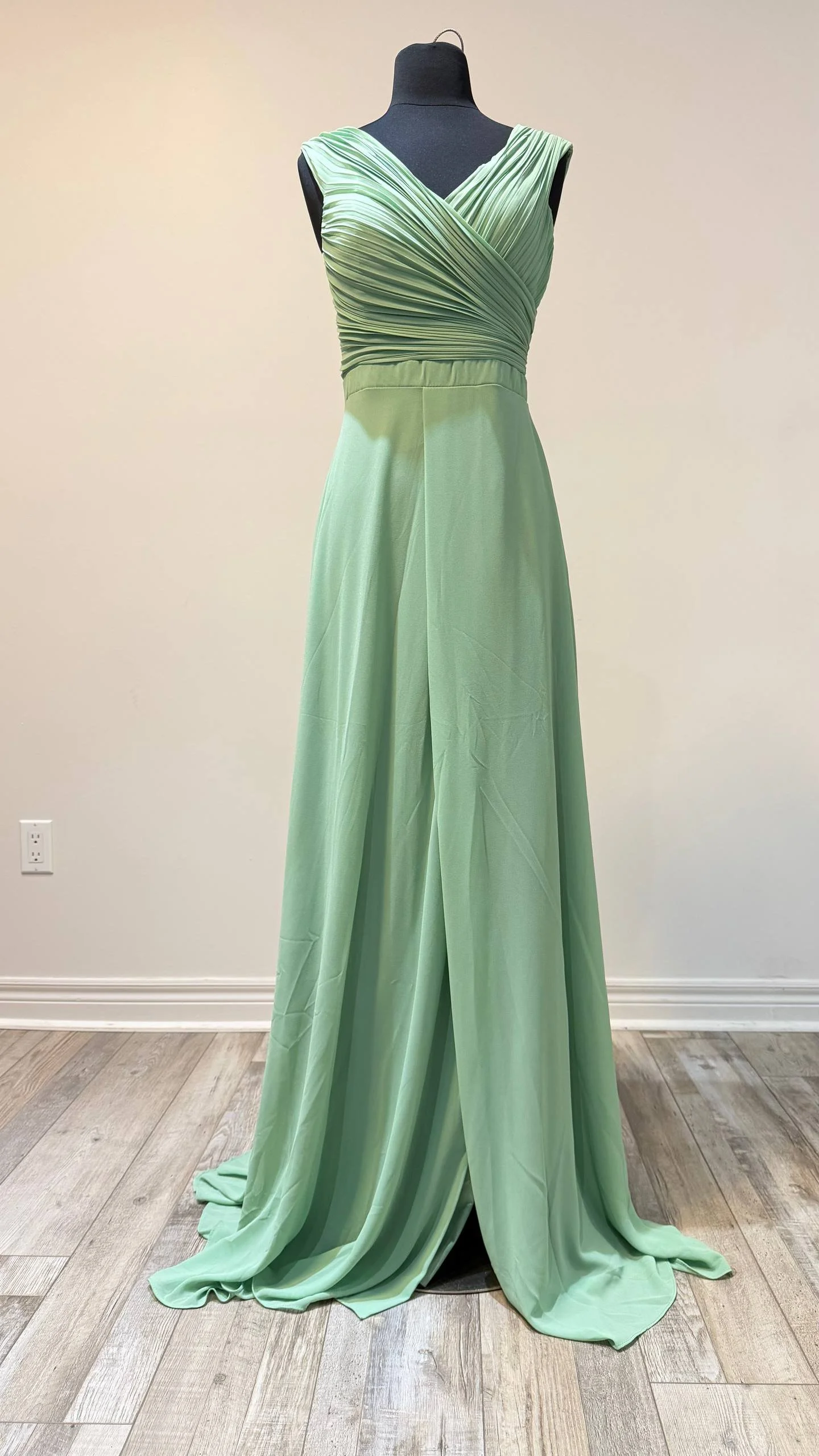 Green evening gown with pleated bodice and flowing skirt displayed on a black mannequin against a plain wall.