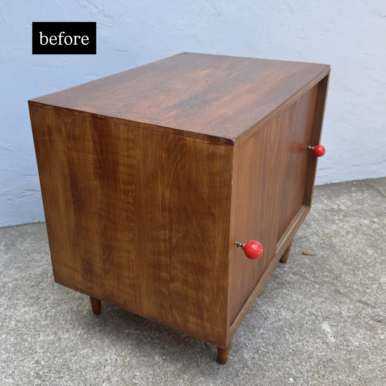 A wooden cabinet with a mid-century modern style, featuring four slim legs and two small round red handles on the side. The cabinet is placed on a concrete surface and is positioned against a light blue wall. The door is closed, and the wood has a wa