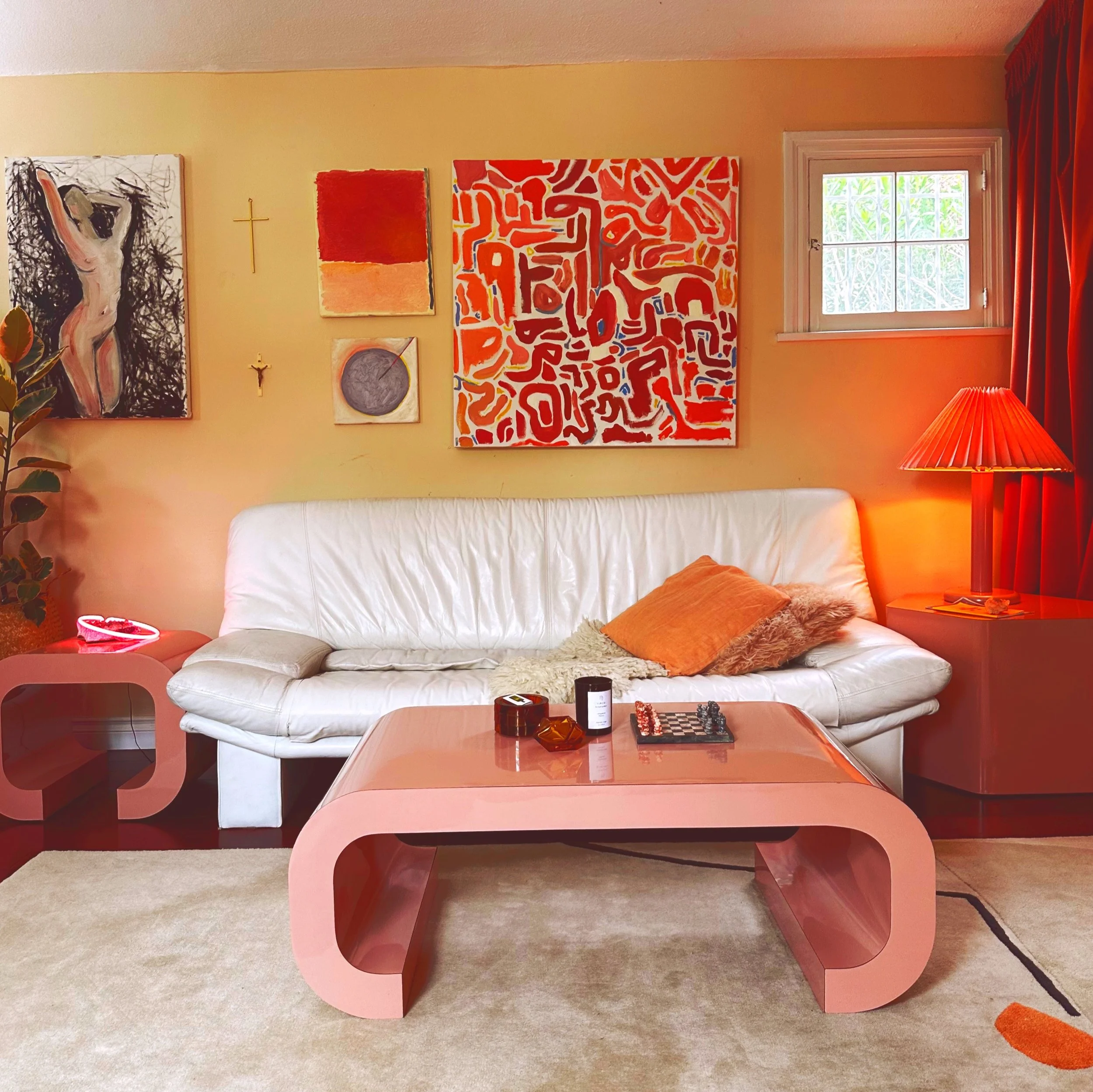 Living room with a white leather couch, pink coffee table, and a pink side table. Wall art includes abstract paintings and a small cross. A window with red curtains and a red lampshade on a side table. Decor includes plants and pillows.