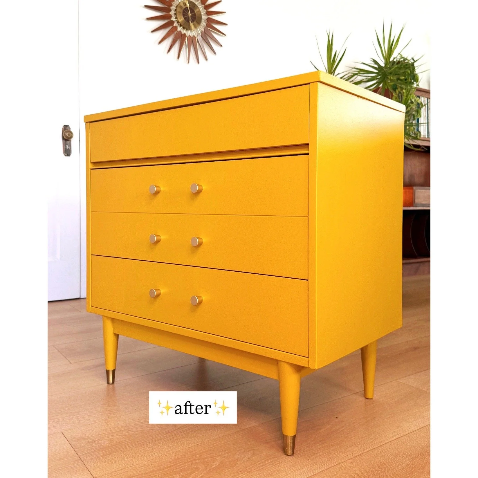 A bright yellow retro-style dresser with four drawers and round knobs, standing on four tapered legs with metallic tips, in a room with light wood flooring and green plants on top, with a decorative wall clock and a white door in the background.