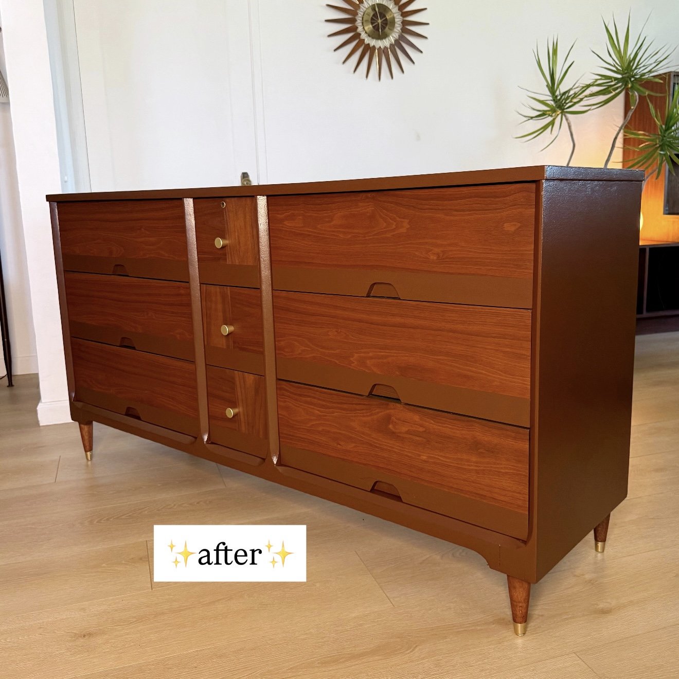A mid-century modern wooden dresser with six drawers and gold knobs, set against a white wall with a sunburst mirror and green plants nearby.