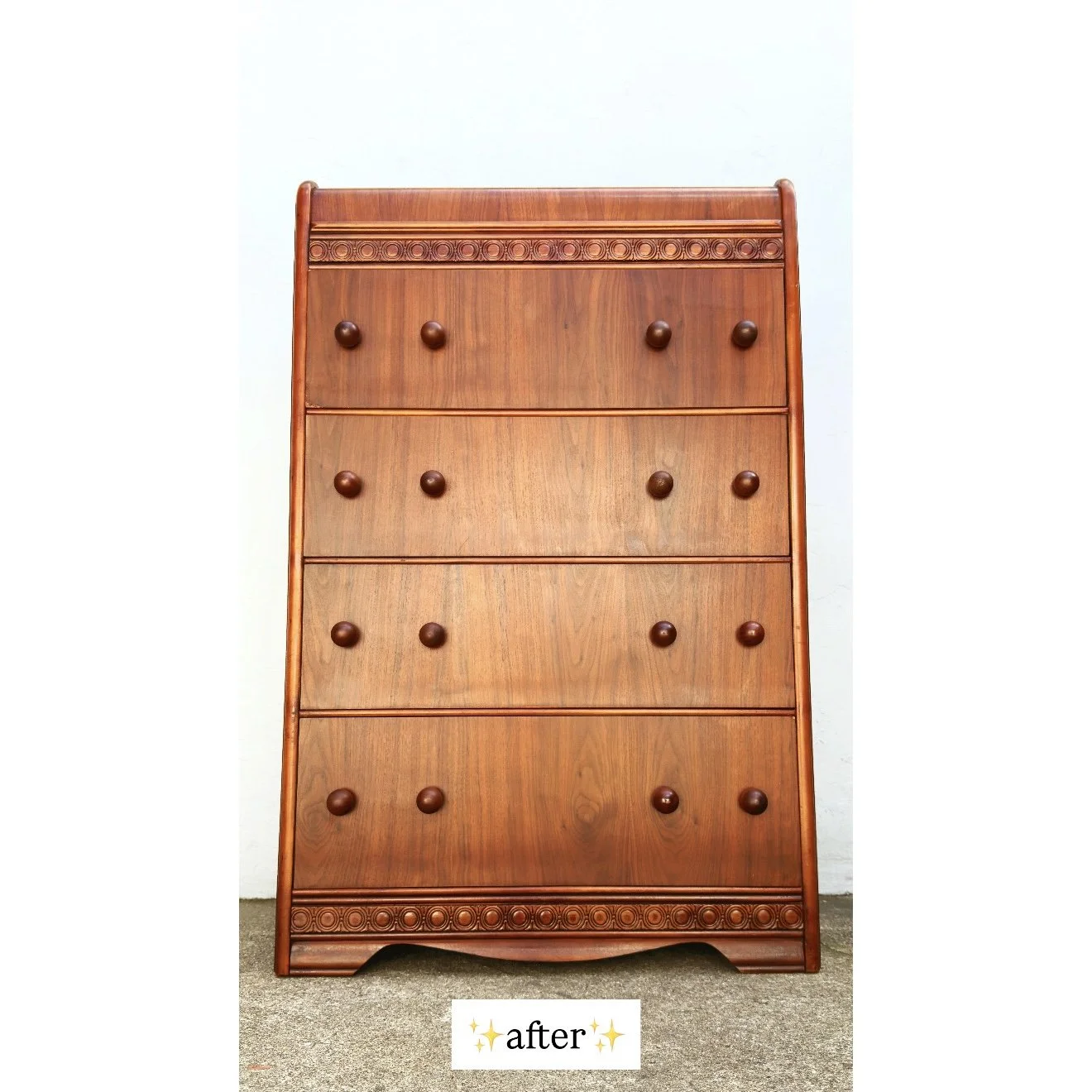 A polished wooden dresser with four drawers and decorative carvings at the top and bottom, set against a white wall with a gray carpeted floor.