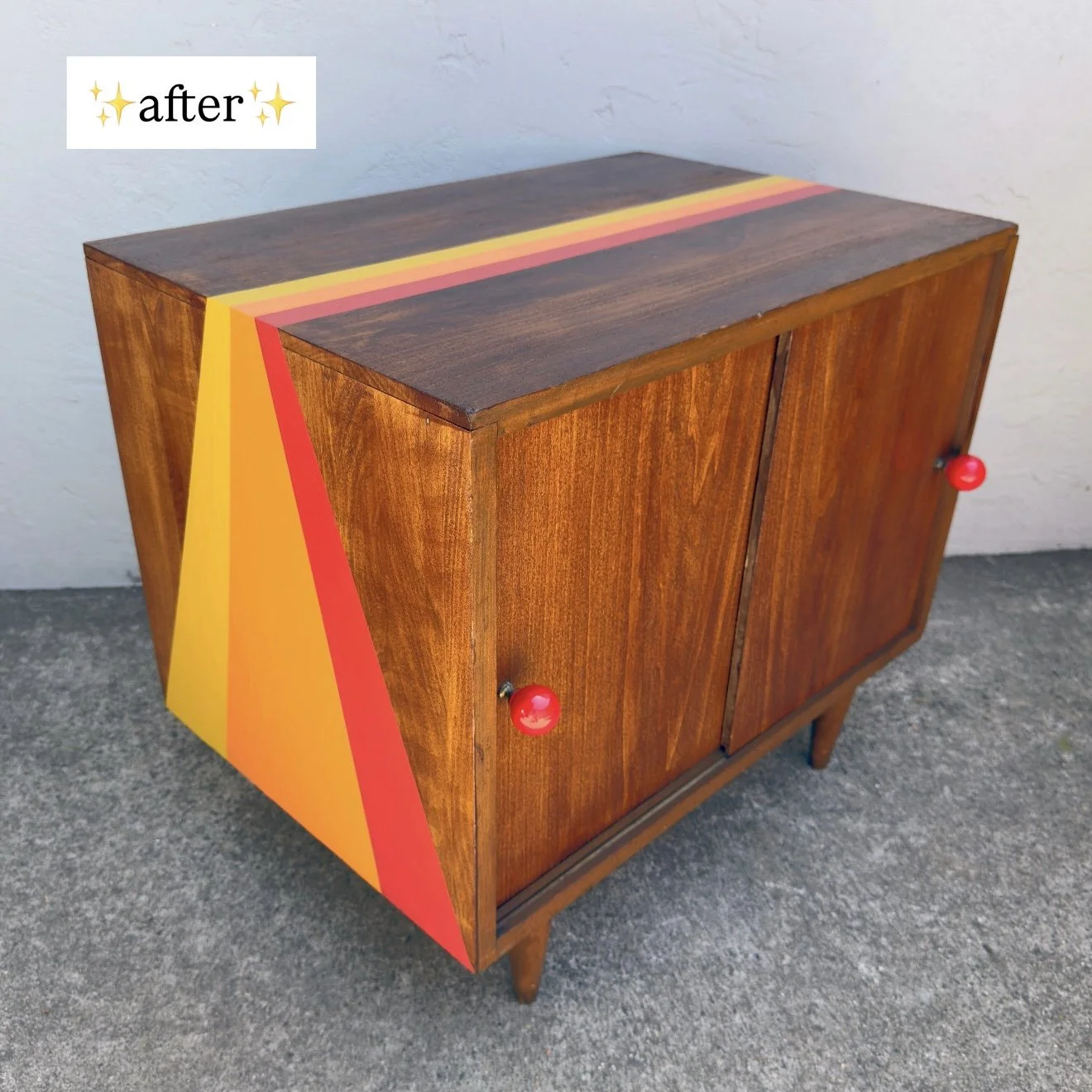 Wooden cabinet with colorful geometric design on one side, featuring yellow, red, and pink stripes, and two red knobs on the doors, placed on a concrete floor against a plain wall with a sticker saying 'after' at the top left corner.