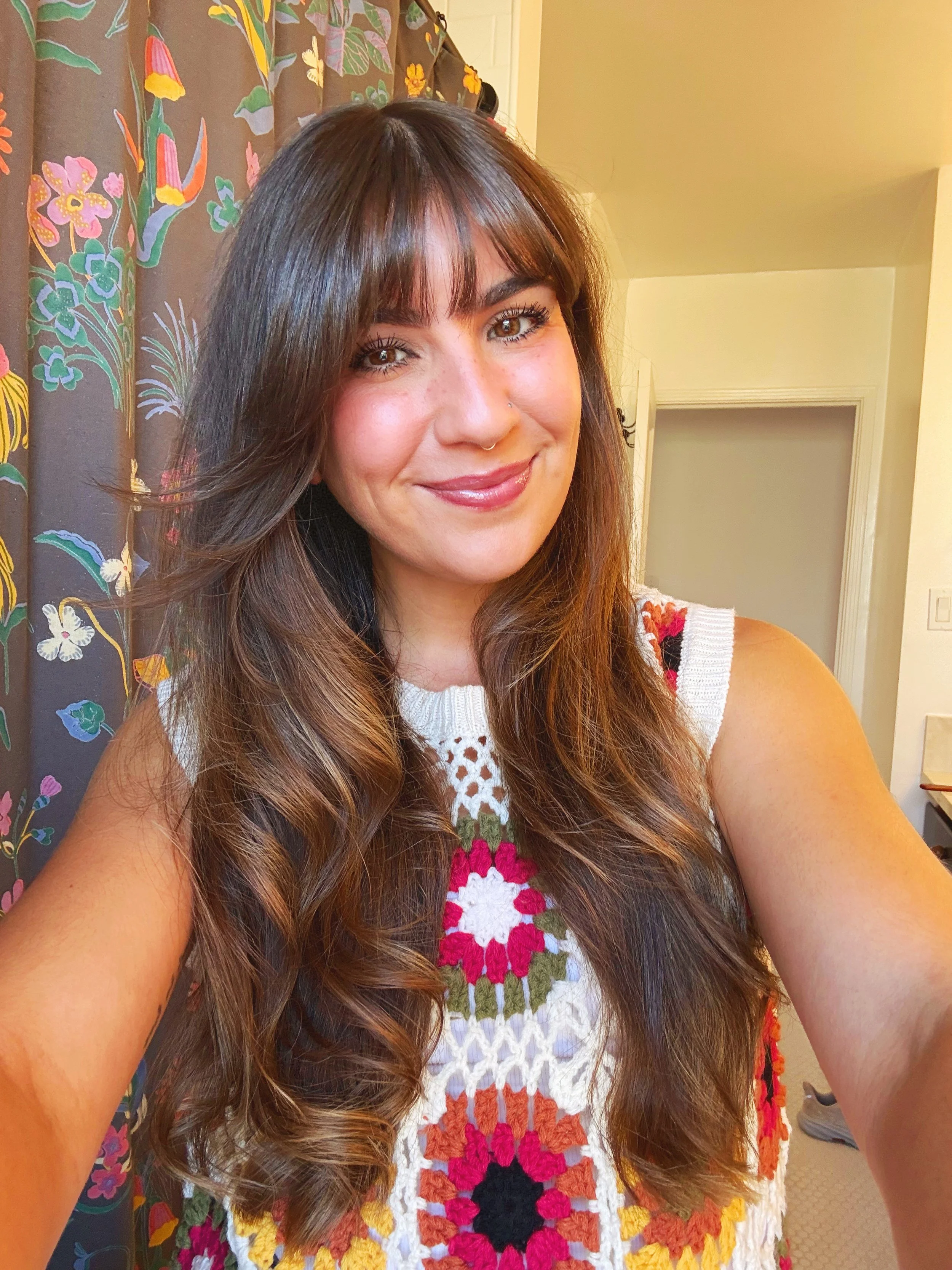 A woman with long wavy brown hair and a nose piercing, smiling at the camera. She is wearing a white crochet top with colorful floral embroidery. In the background, there is a floral curtain with pink, yellow, and purple flowers.