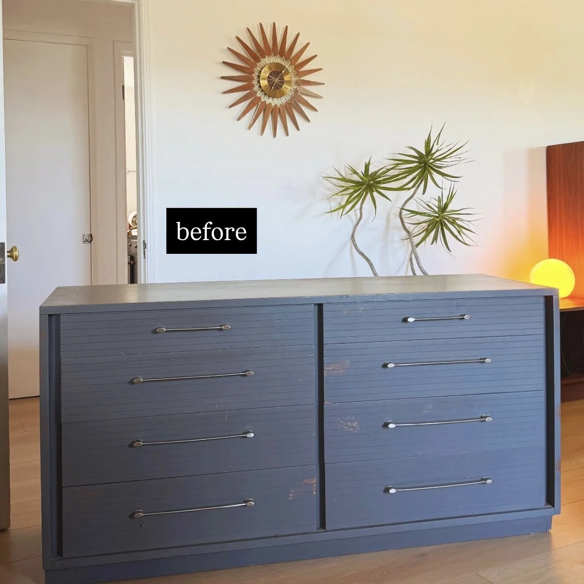 A room with a mid-century modern blue dresser, a wall clock with a sunburst design, a potted plant with long, spiky green leaves, and a yellow glowing lamp on a wooden side table, indicating the space is shown before a makeover.