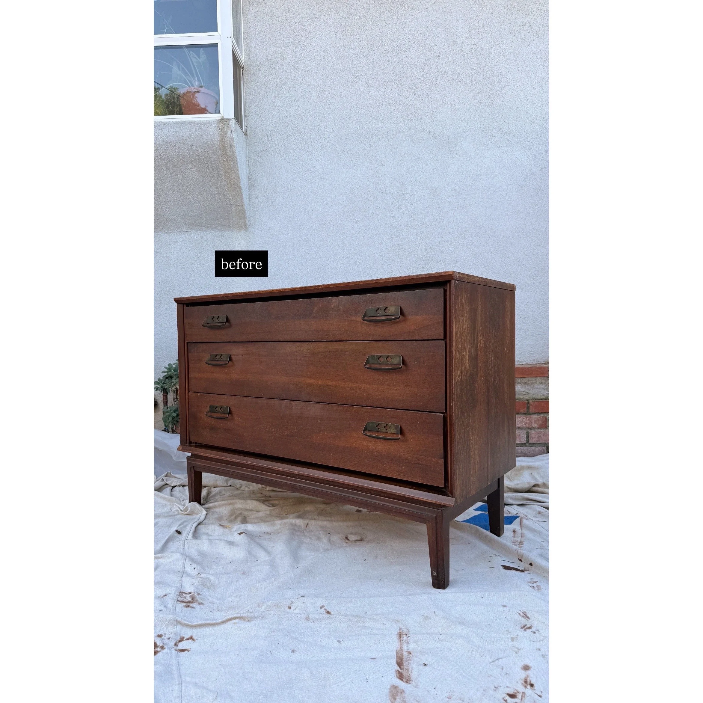 Wooden dresser with four drawers and brass handles on a white cloth outdoors against a light gray wall with a window in the corner.