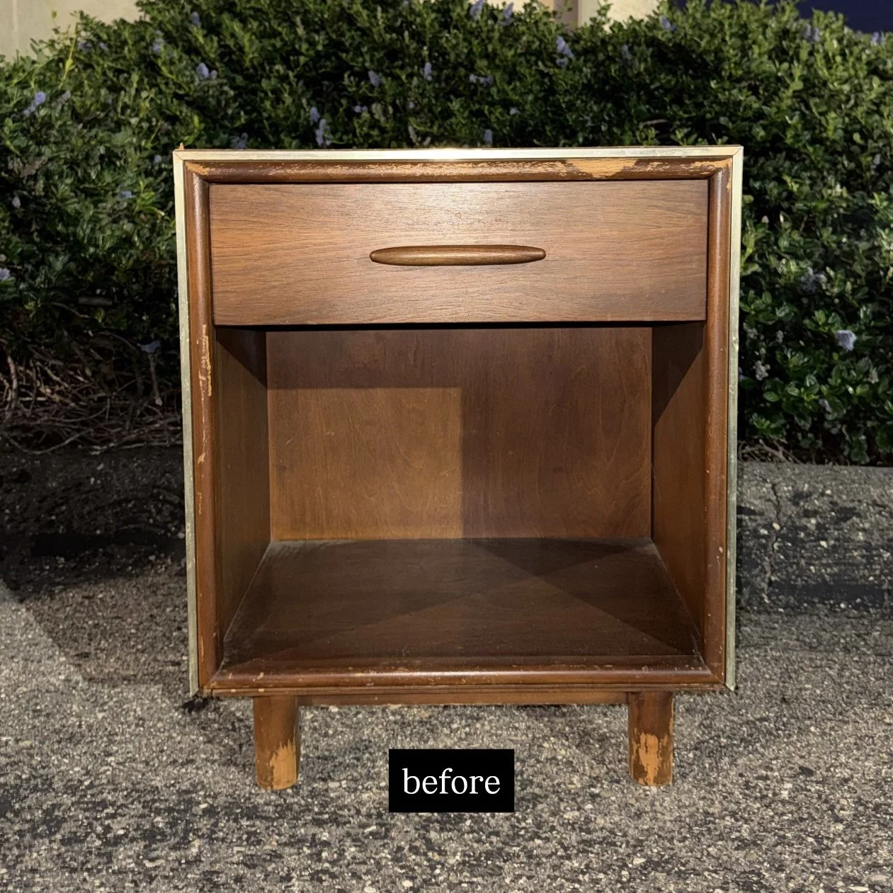A vintage wooden nightstand with a drawer at the top, showing signs of wear and chipping on the legs and edges, placed on asphalt with greenery in the background.