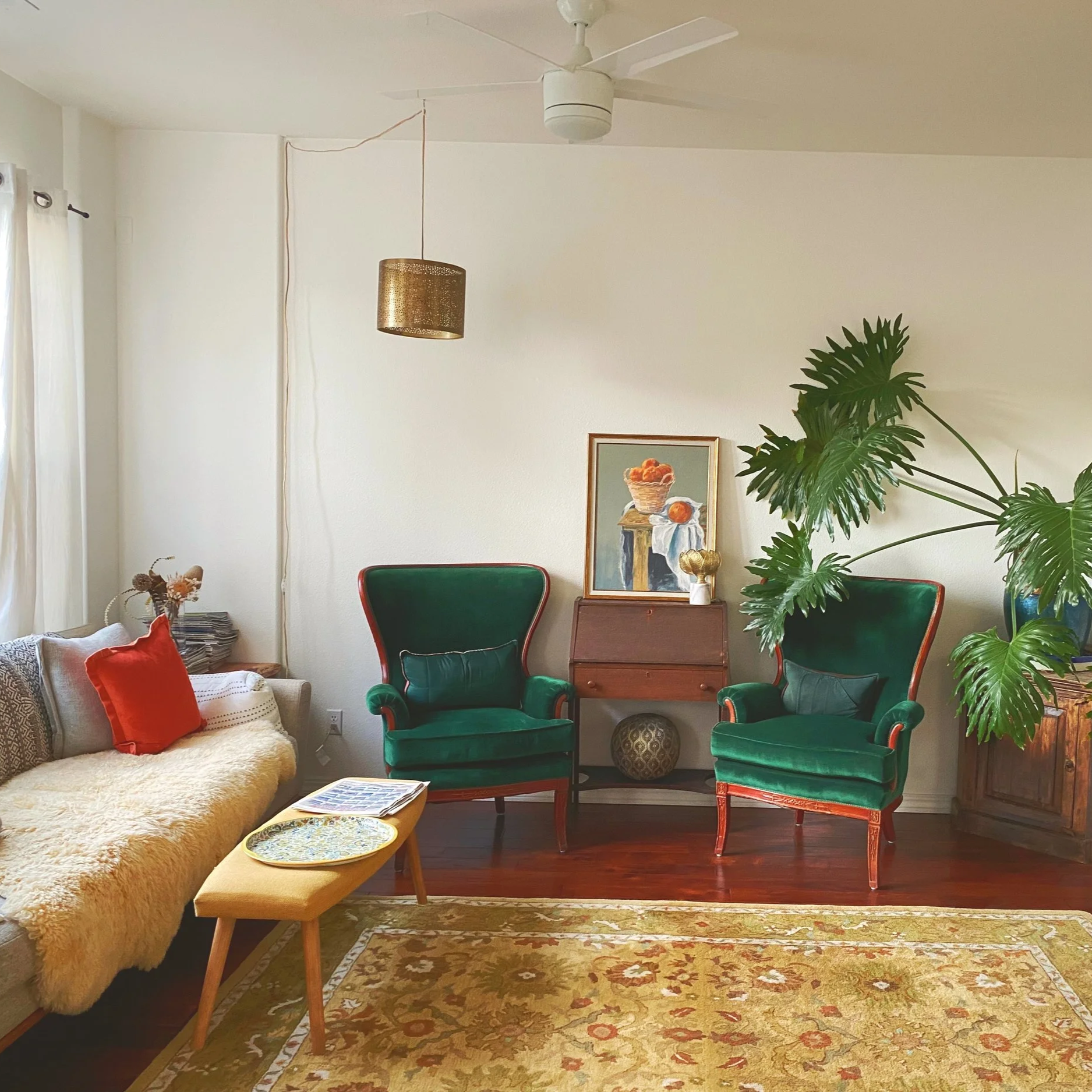 Living room with a beige sofa, red pillow, green armchairs, a wooden side table with art, a large green plant, a ceiling fan, and a patterned rug.