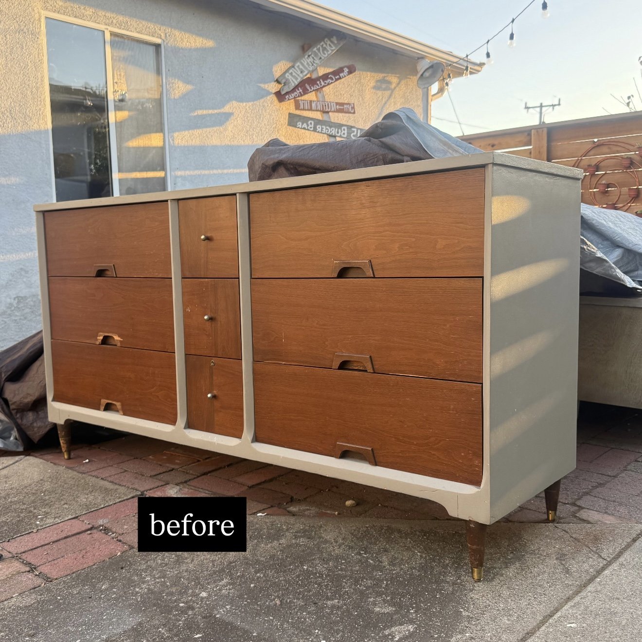 A vintage wooden dresser with multiple drawers and a white frame, set outdoors on a brick and concrete surface, with a house, window, and outdoor lighting in the background.