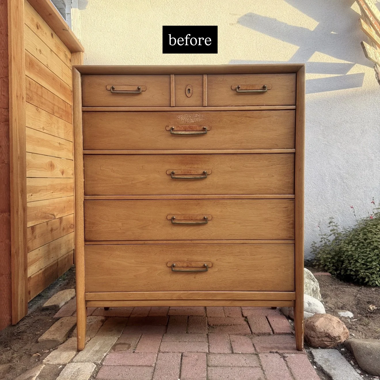 A wooden dresser with six drawers standing outside on a brick-paved area near a white wall and a wooden fence, with a label saying 'before' at the top.