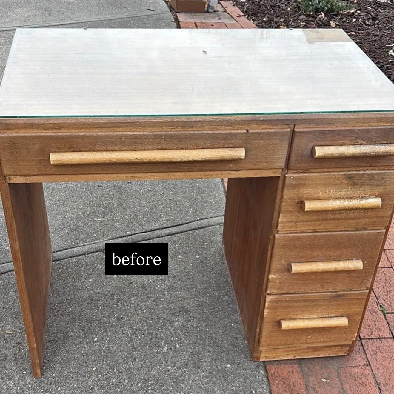A wooden desk with a glass top, positioned outdoors on a concrete surface. The desk has a drawer on the left side with a handle, and four drawers on the right side, each with handles. There is a small label reading 'before' on the concrete near the l