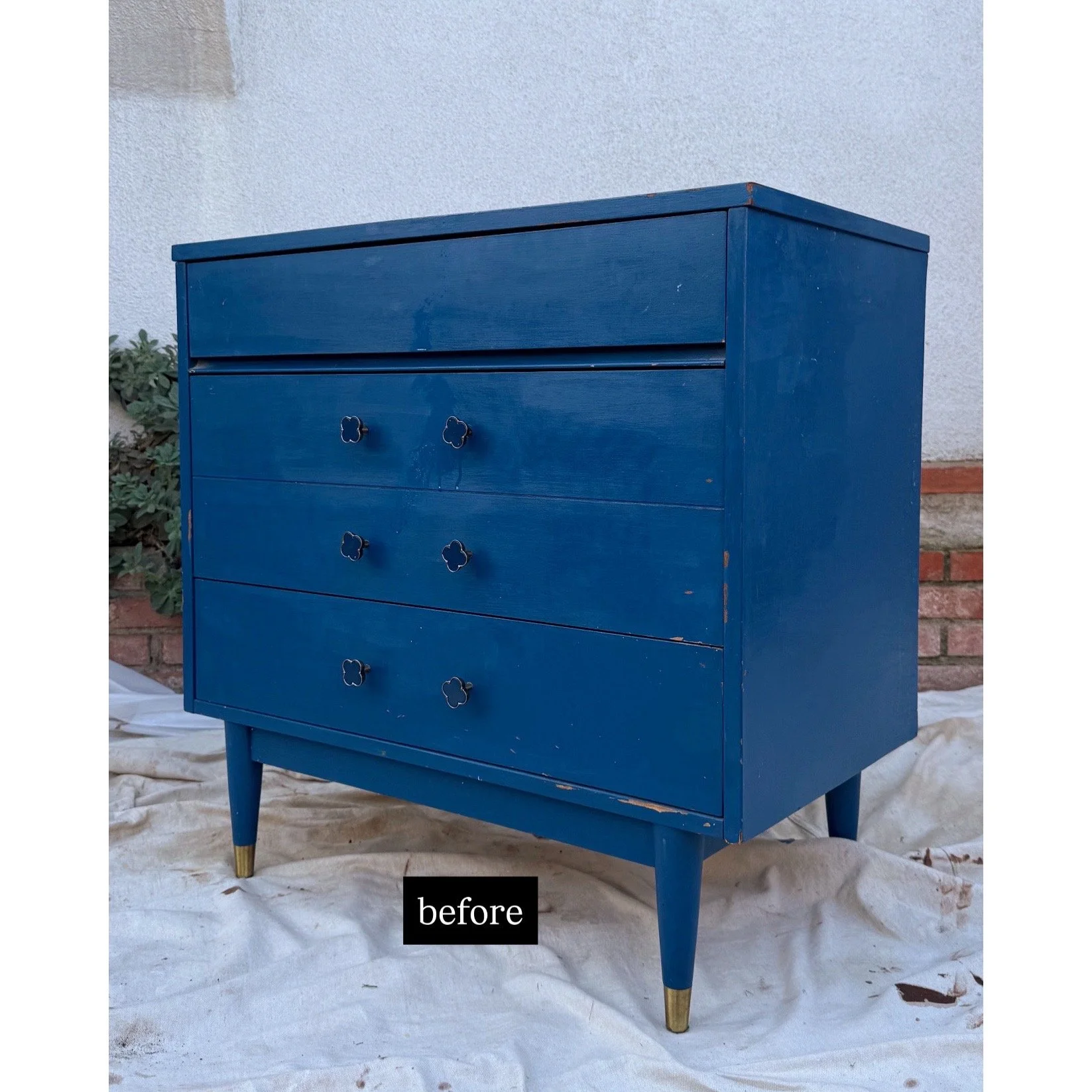 A vintage blue dresser with four drawers and gold-tipped legs, placed outdoors on a white cloth, against a white wall with some brick details and greenery.