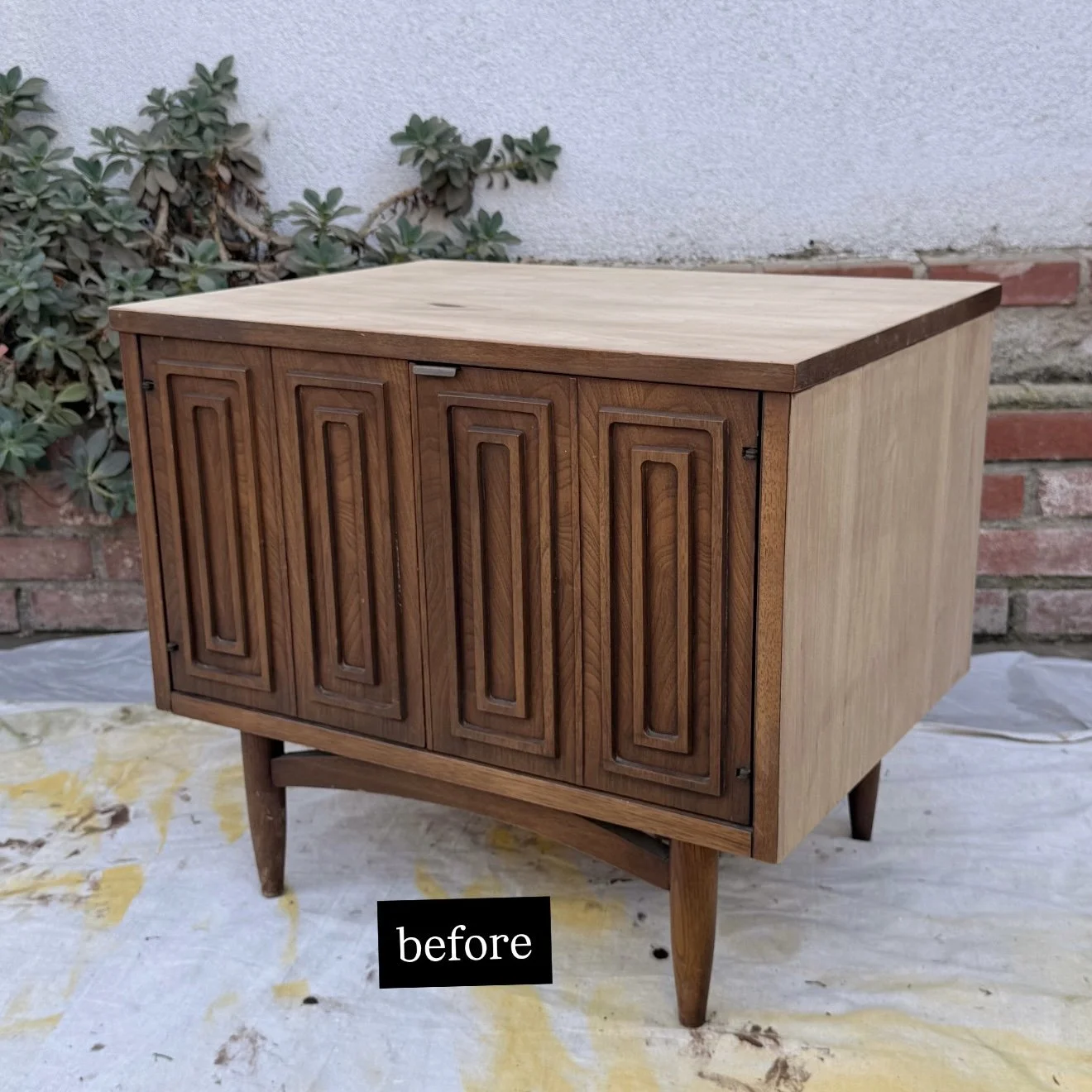 A vintage wooden cabinet with four paneled doors and tapered legs, placed outdoors on a tarp with a brick wall and plants in the background.