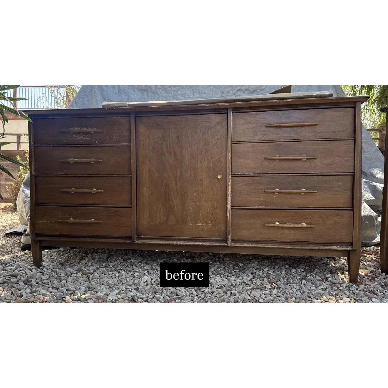 Old wooden dresser with multiple drawers and a sliding door, sitting outdoors on a gravel surface.