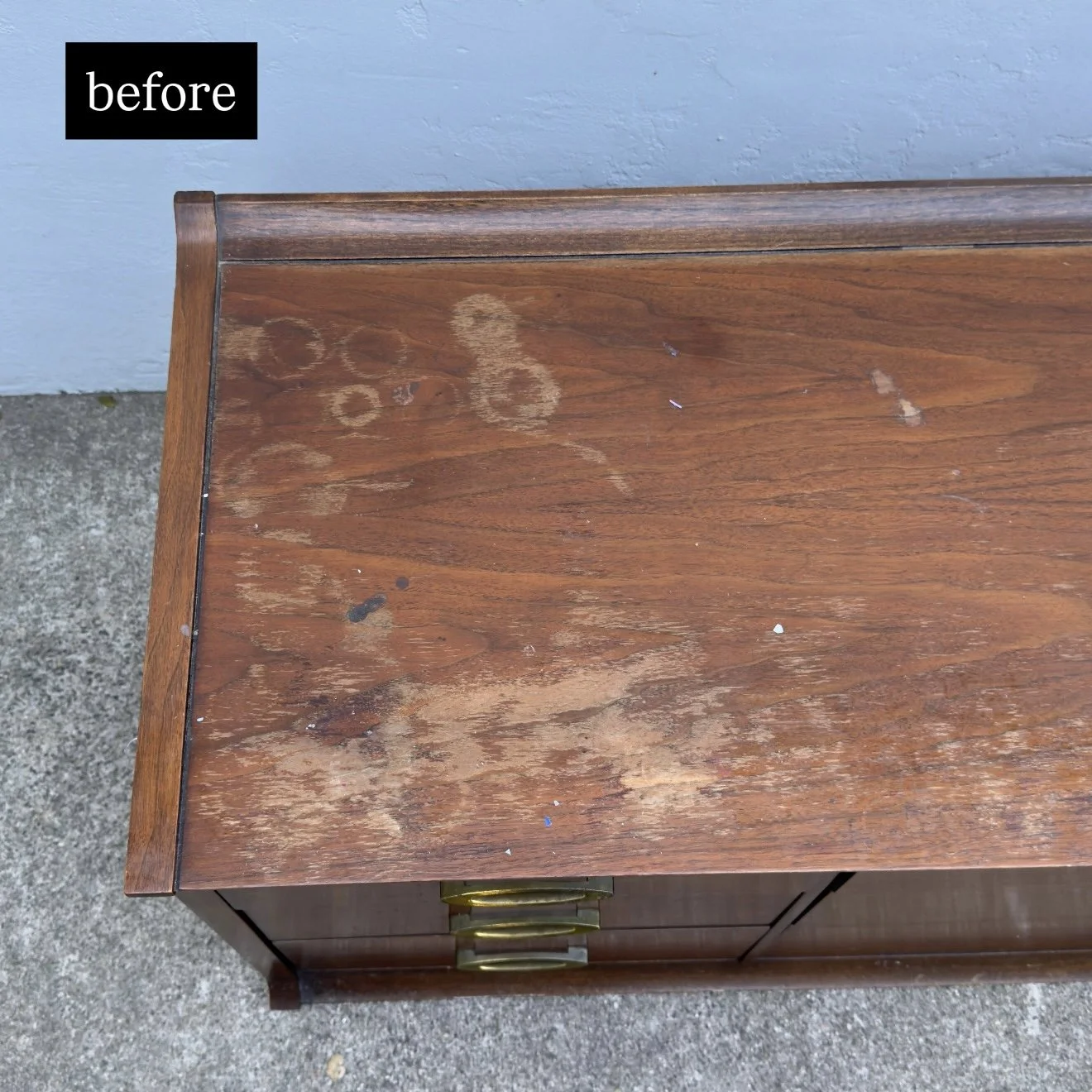 Wooden top table with scratches, stains, and wear, placed on concrete, with a blue wall in the background.