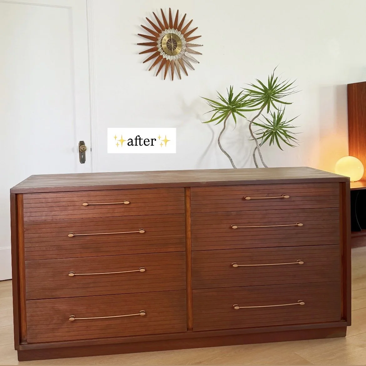 A mid-century modern wooden dresser with horizontal grain and round handles, positioned against a white wall with a decorative sunburst clock and a potted plant with long, branch-like stems. A white orb-shaped lamp is in the background to the right.