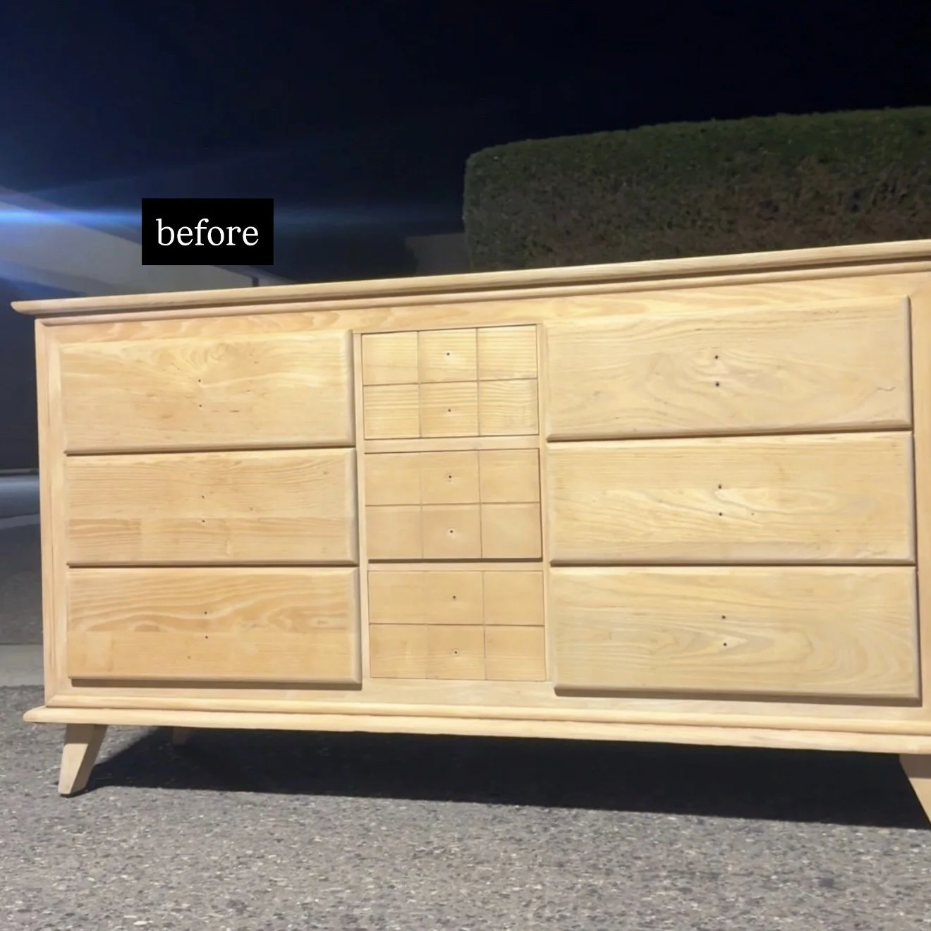 A wooden dresser with multiple drawers, shown before refinishing or staining, is set outside at night on a paved surface with a hedge in the background.