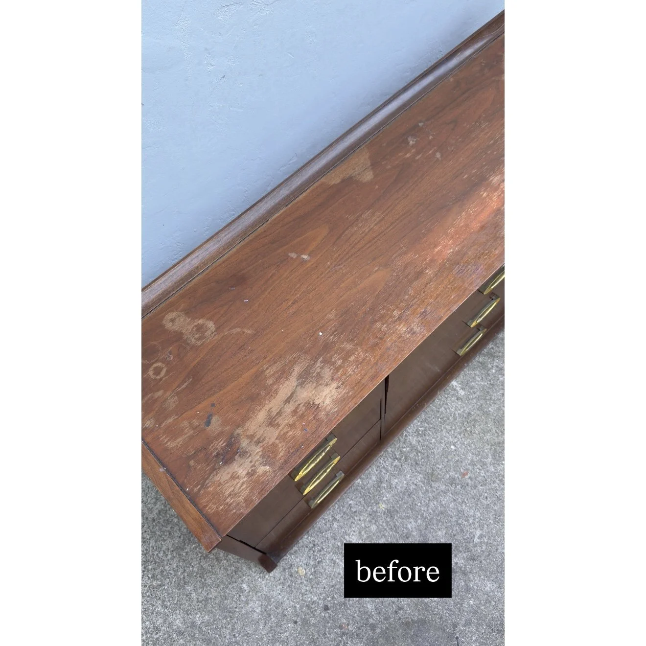 A wooden dresser with a scratched and worn top surface, showing areas of discoloration and damage, placed against a textured wall and sitting on a concrete floor.