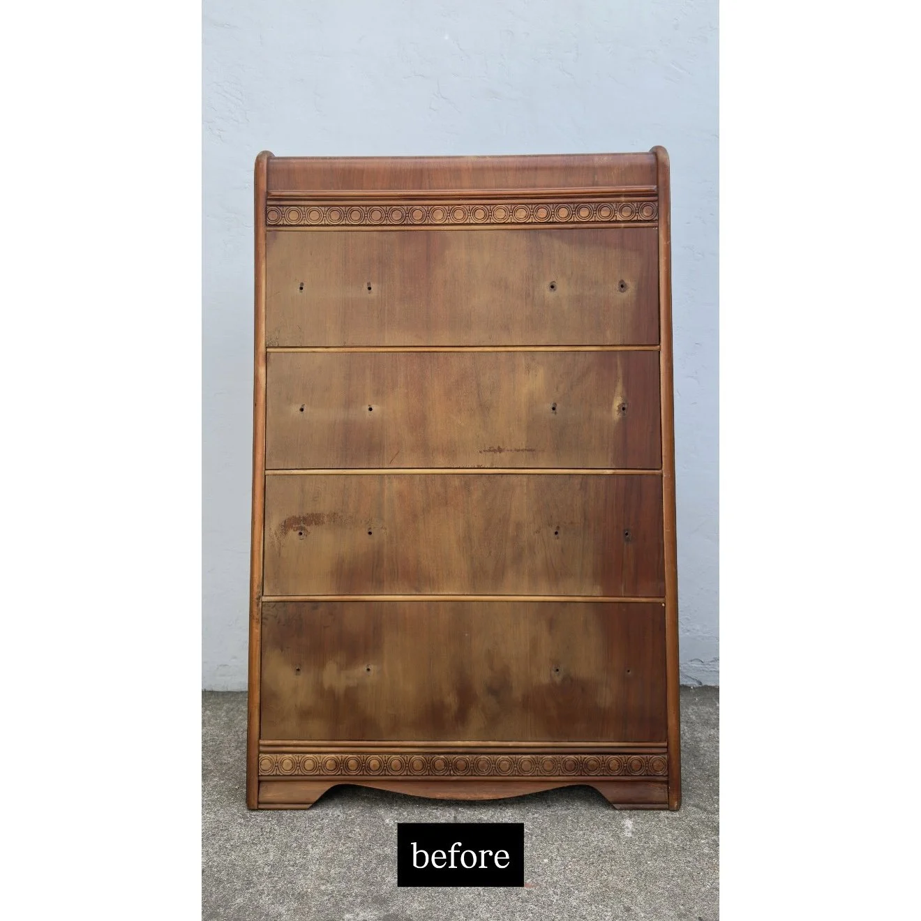 A wooden headboard with carved decorative details at the top and bottom, leaning against a light-colored wall on a gray carpeted floor.