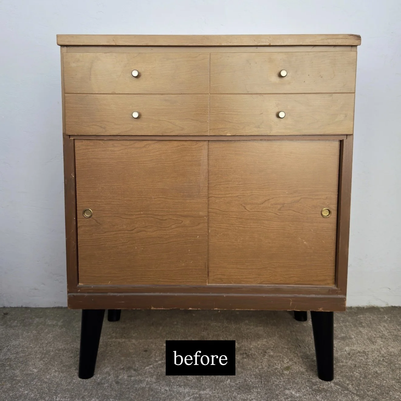 An old wooden cabinet with four drawers and sliding doors, showing worn edges and scratches, set against a light gray wall.