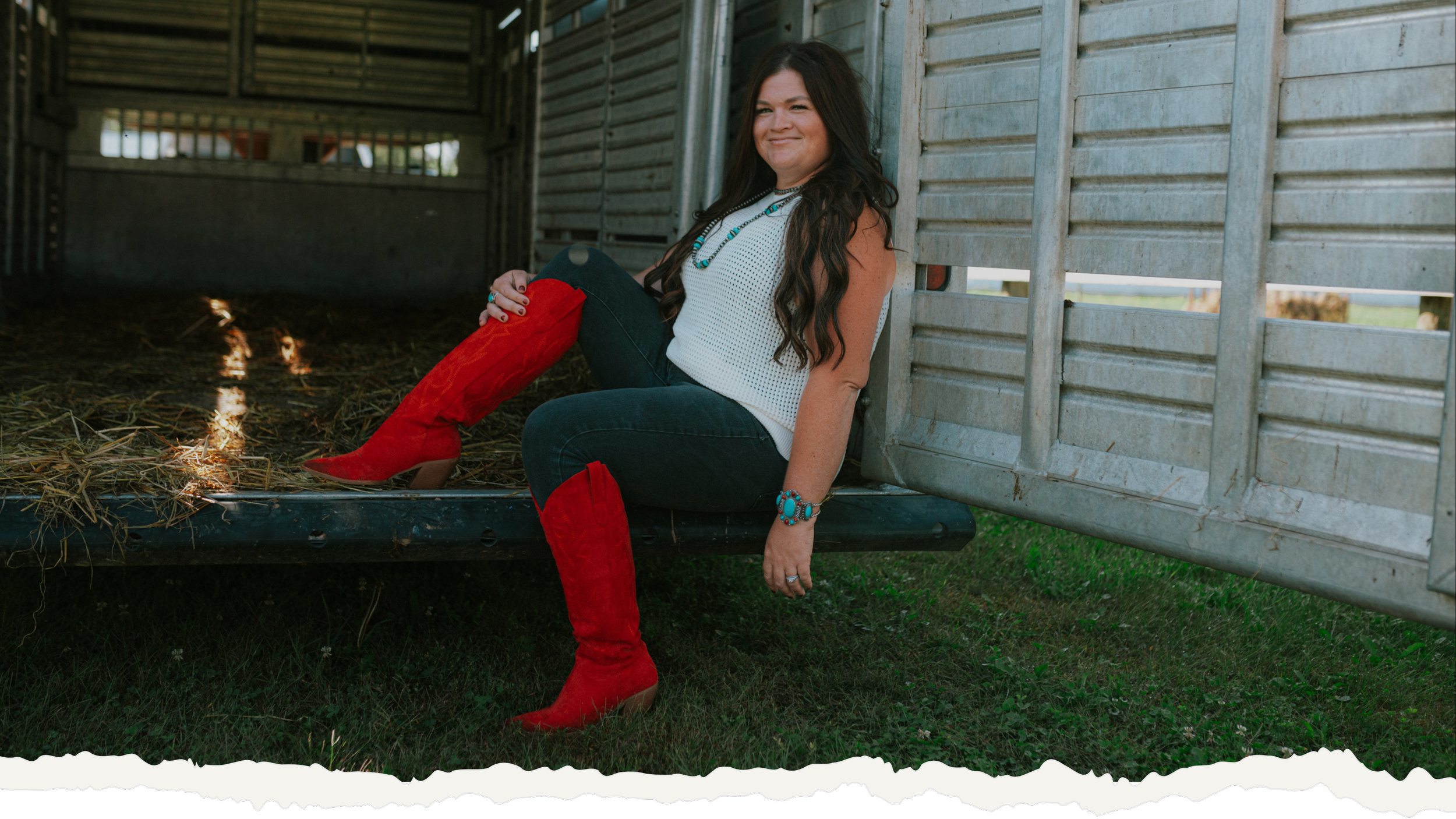 Ryann sitting on the edge of a livestock trailer, smiling, wearing a sleeveless white top, dark jeans, and red cowboy boots, with turquoise jewelry.