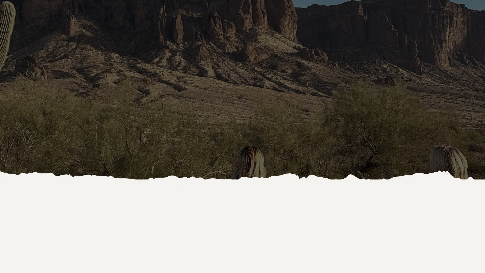 A desert landscape with rocky mountains, sparse vegetation, including bushes and cacti, with two women with blonde hair in the foreground.