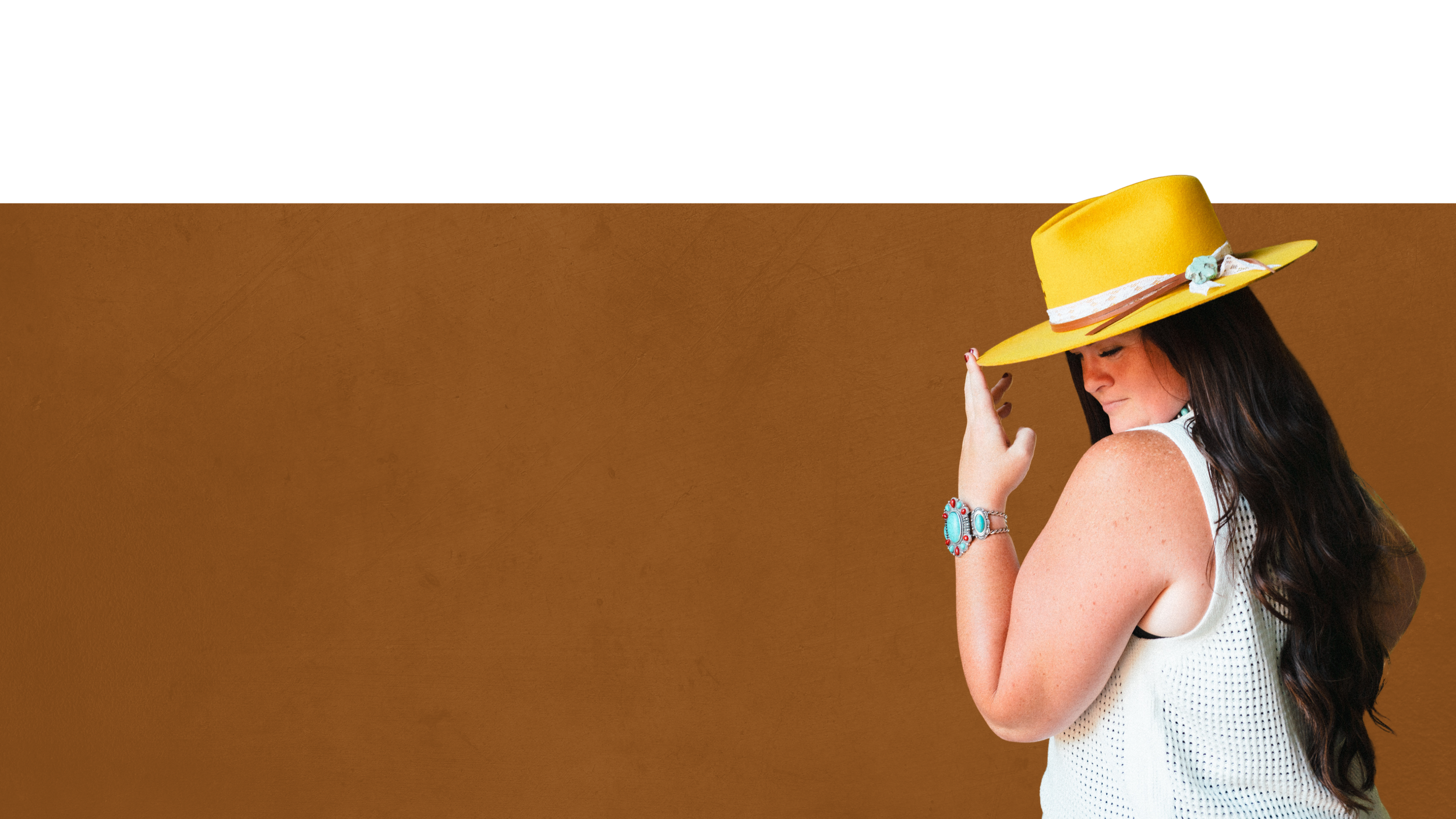 Ryann in a yellow wide-brim hat with turquoise jewelry smiling with eyes closed, wearing a white sleeveless top against a warm brown backdrop