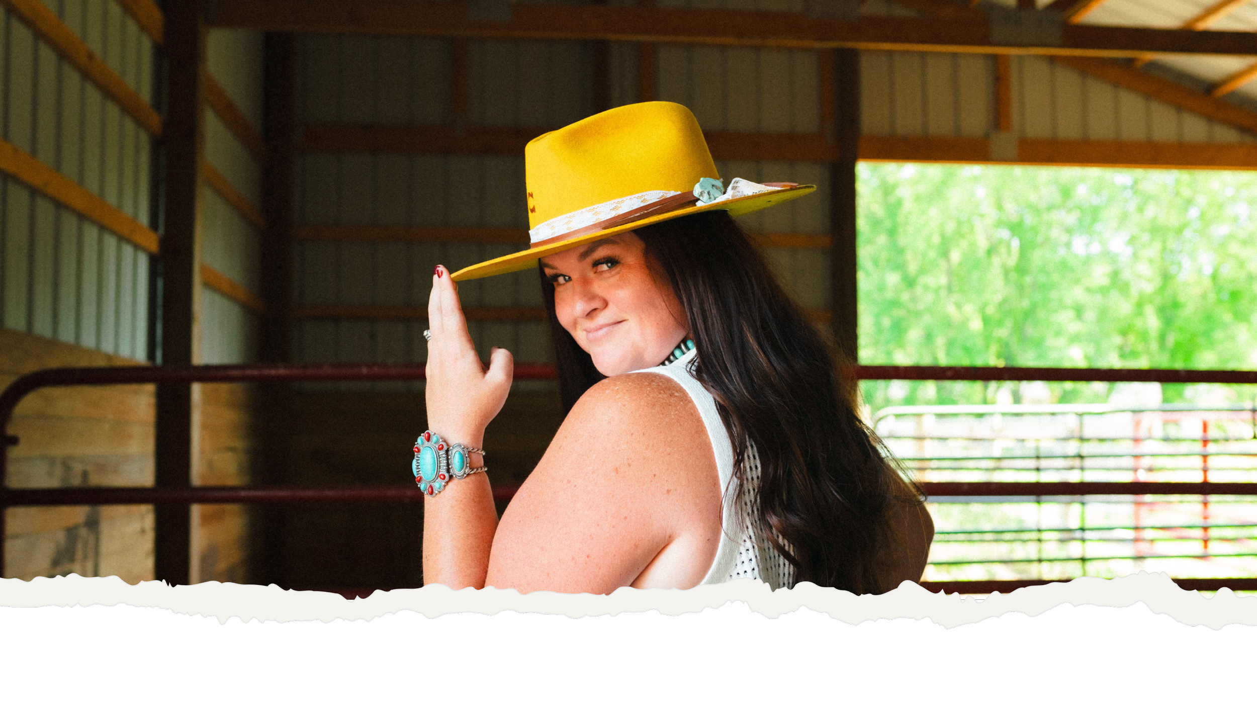 Ryann with long dark hair, wearing a yellow hat and a white sleeveless top, is smiling and looking over her shoulder inside a barn or stable with green trees visible outside.