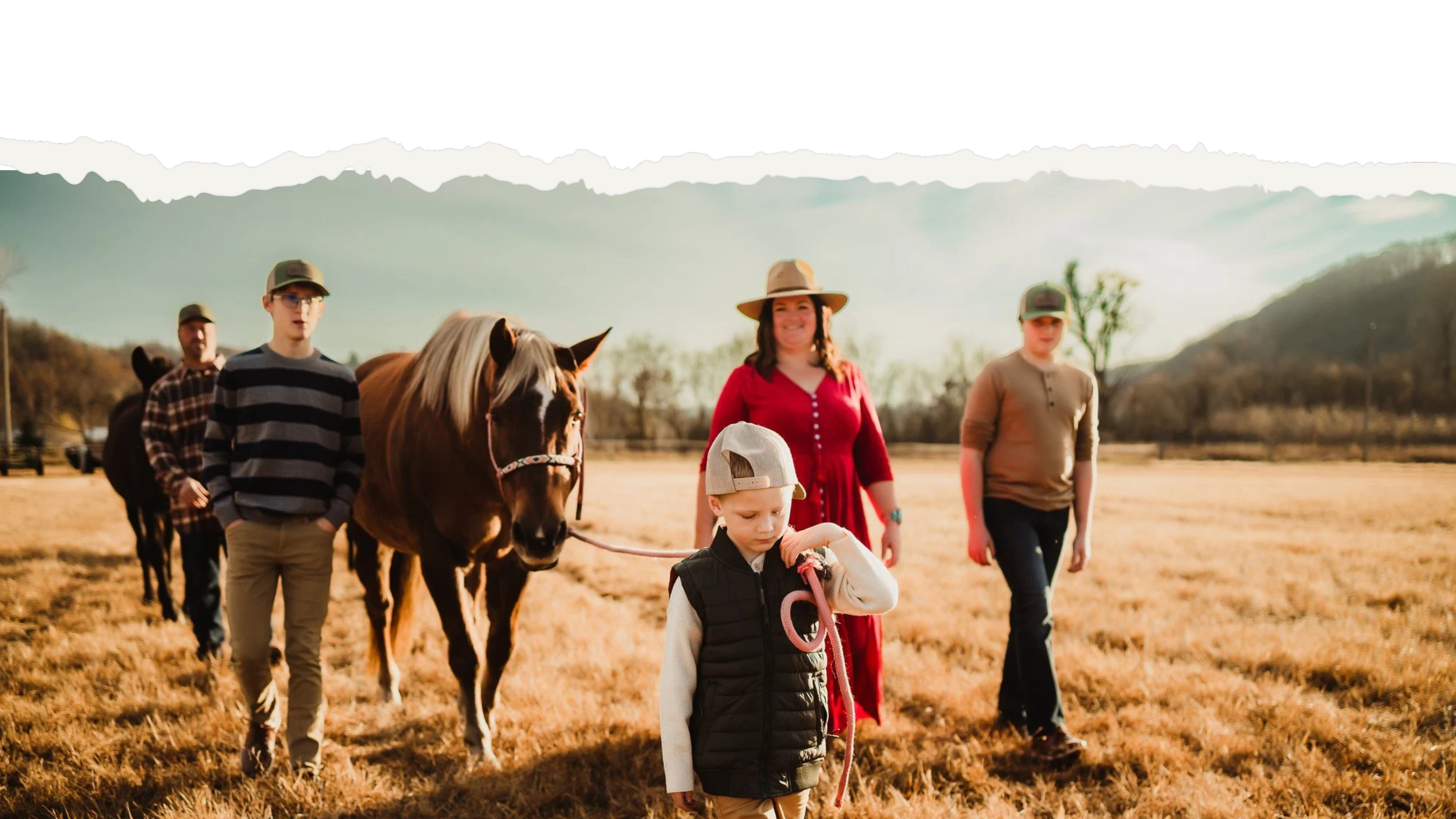 Ryann and family walking outdoors in a field with mountains in the background, including a woman in red, three children, and a person leading a horse.