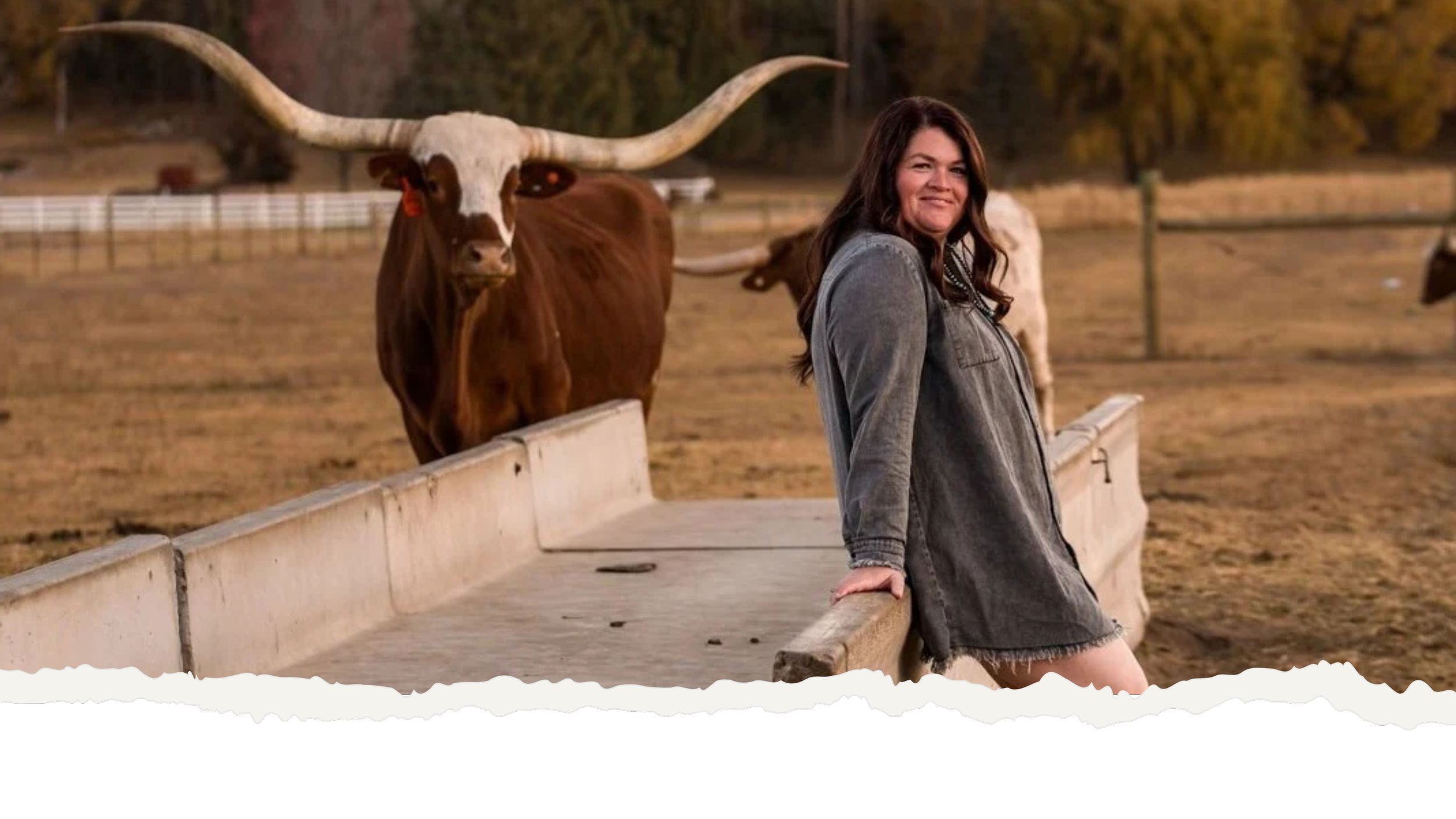 Ryann in a gray jacket smiling and leaning on a wooden fence at a farm with a large longhorn steer with prominent horns behind her.
