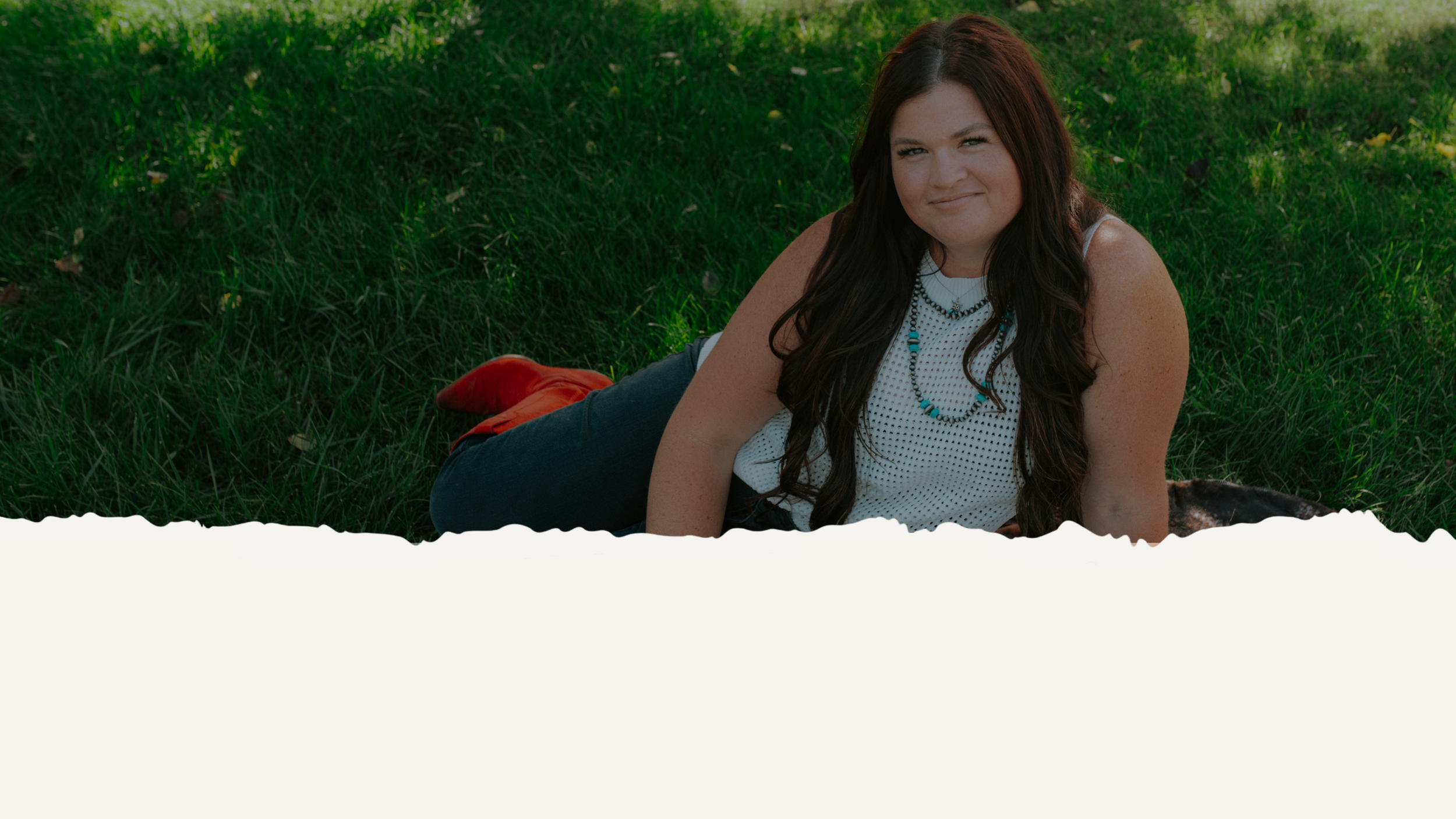 Ryann Bauer with long dark hair lying on grass in a park, wearing a white sleeveless top, dark jeans, red shoes, and layered necklaces, smiling at the camera.