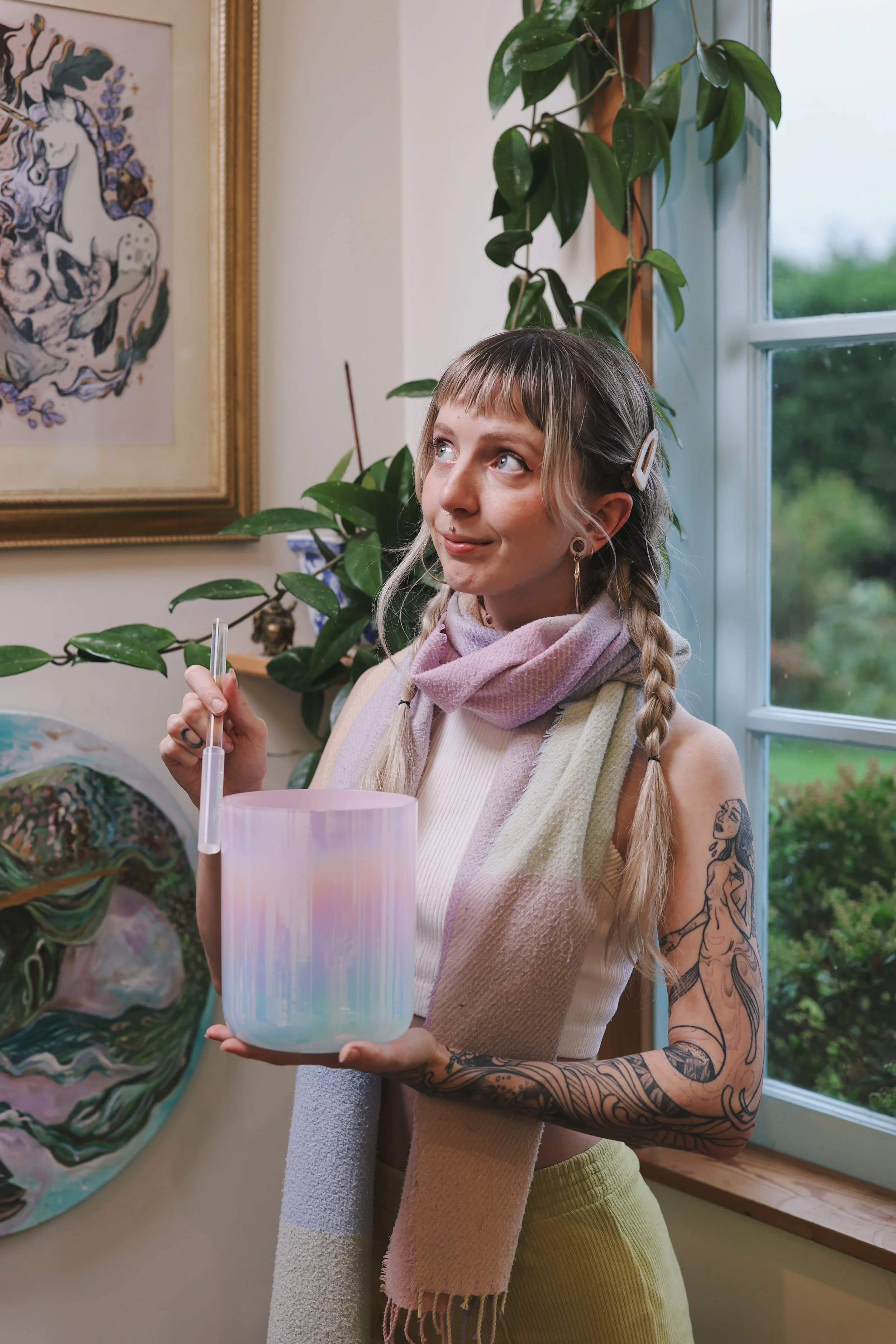 Young woman with tattoos on her arm, holding a colorful crystal singing bowl, standing indoors near a window with a green outdoor view, and artwork on the wall behind her.