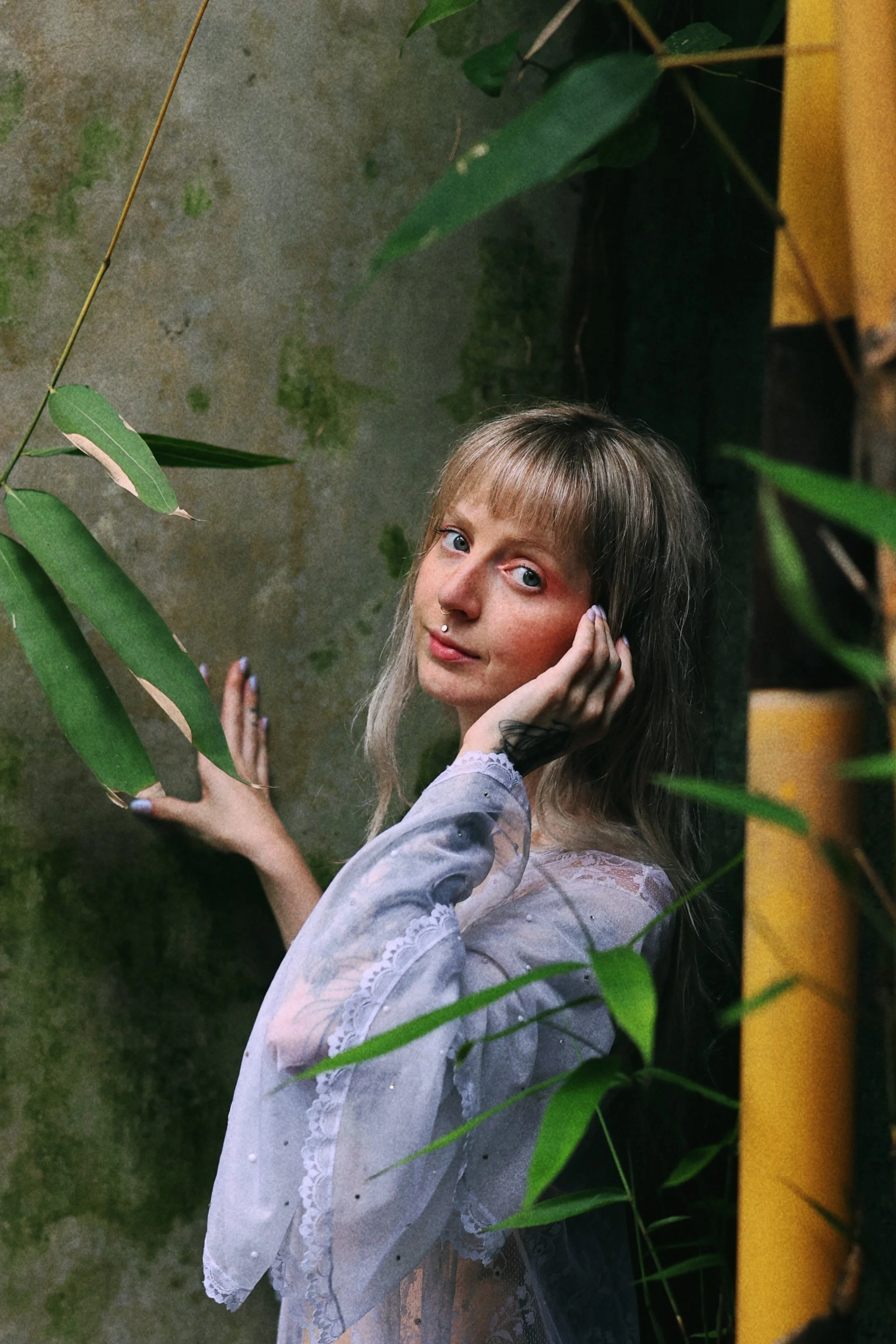 A young woman with a patterned headscarf and layered clothing sits at a wooden table with tarot cards fanned out in front of her. There is a small vase with dried flowers on the table, and a pink tufted wall in the background.