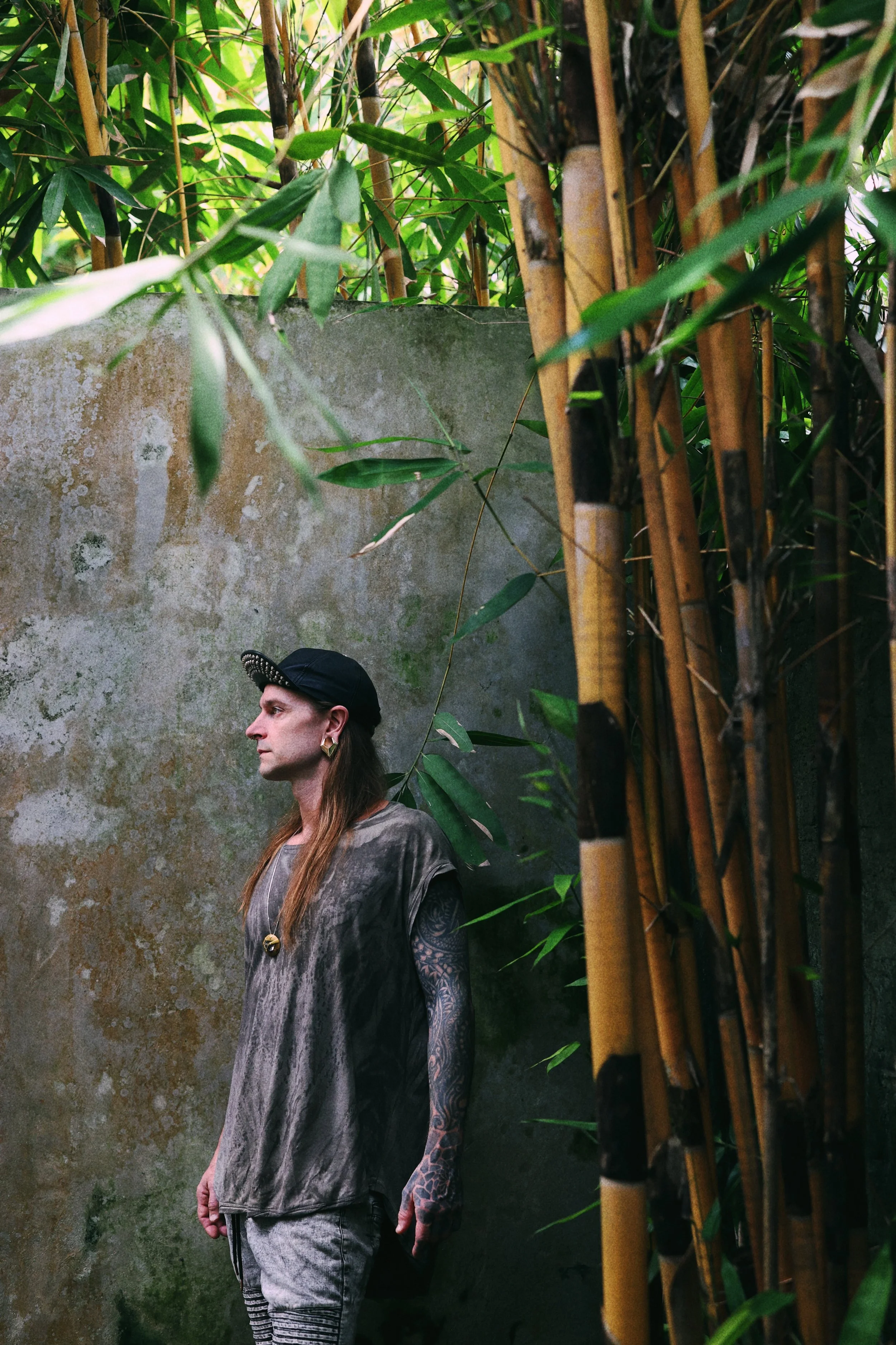 A man with long hair, earrings, a nose ring, and tattoos posing outdoors with a house and blooming bushes in the background.