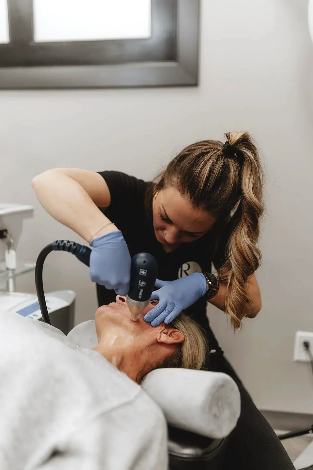 A woman in gloves performing a skincare procedure with a handheld device on a woman lying down, in a clinical setting.