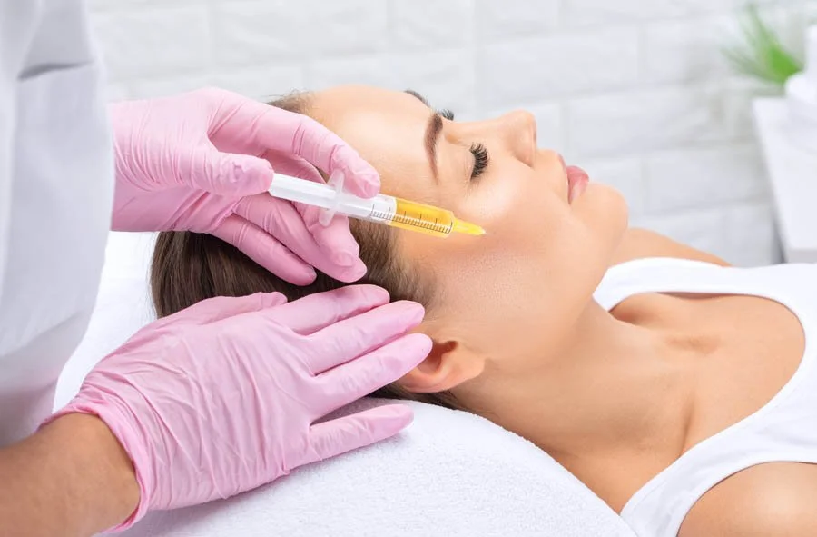 A woman is receiving a facial treatment at a skincare clinic, with the esthetician applying a product to her forehead. The woman has a headband and a facial mask on. The setting is modern with a sign that says "The Refinery" on the wall.